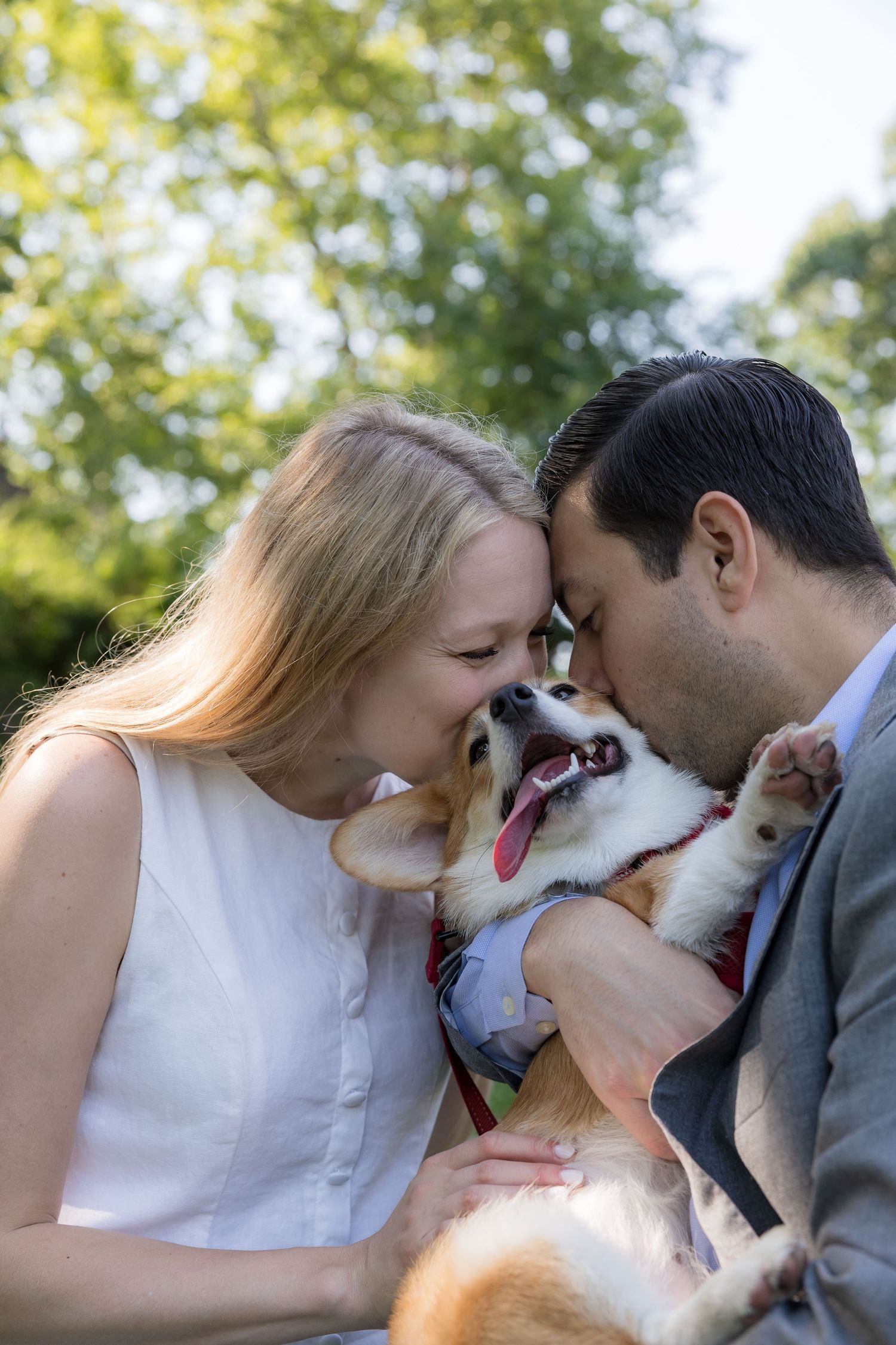 Happy couple kissing their smiling Corgi dog outdoors with green trees in the background.