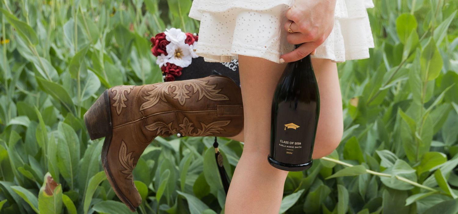 Close-up of cowboy boots and wine bottle held by woman in white dress in corn field.