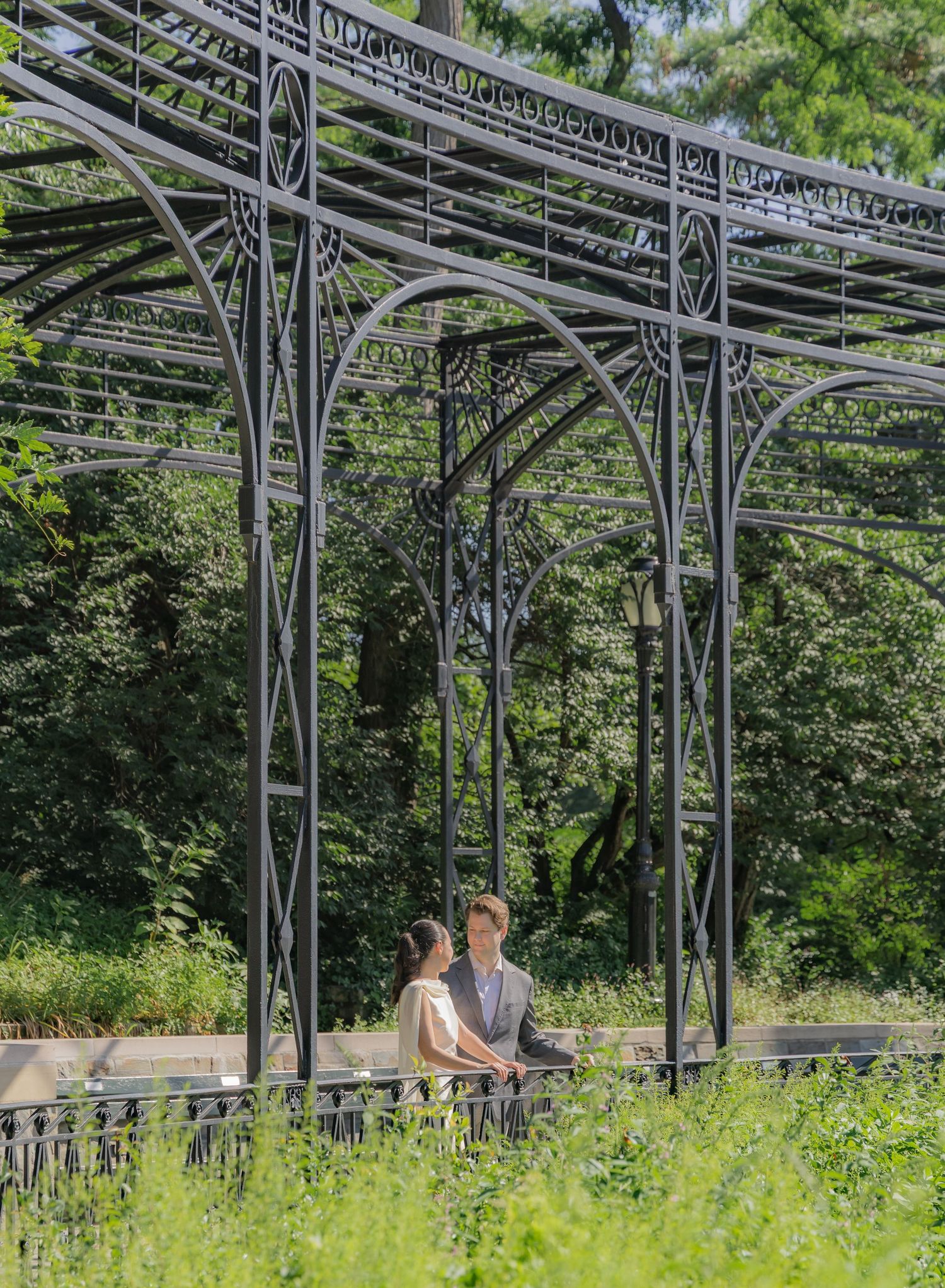 Couple sitting together under an ornate black iron pergola surrounded by lush green trees in a garden setting.