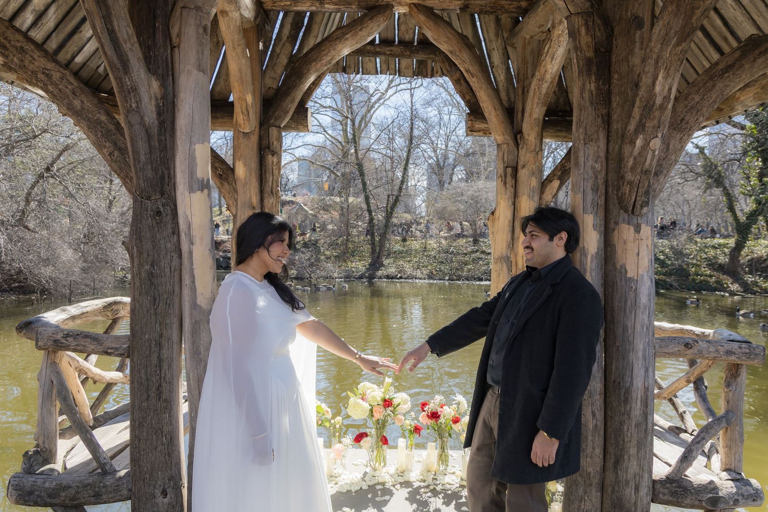 Bride and groom hold hands under rustic wooden gazebo in Central Park, with a colorful bouquet and pond in background.