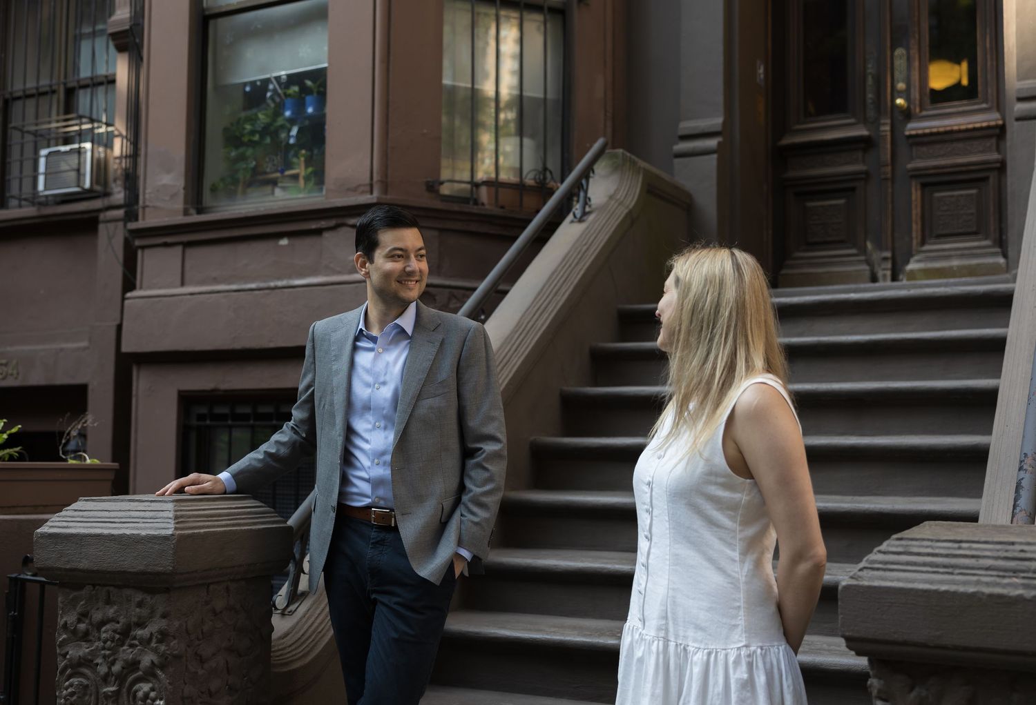Couple smiling at each other outside a NYC brownstone, man in blazer holding hat, woman in white dress.