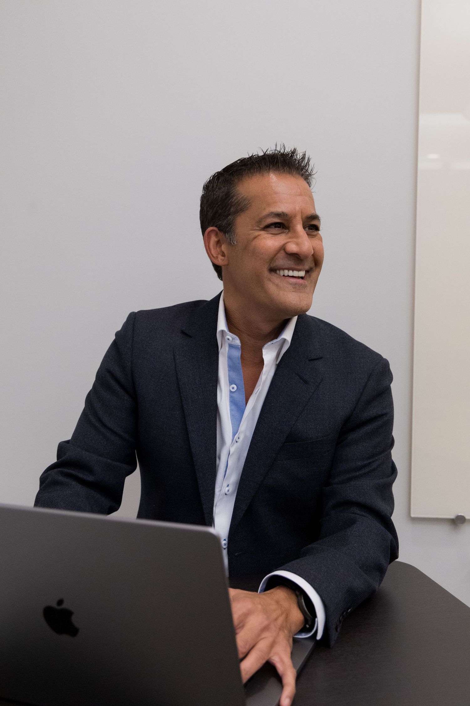 A businessman in a dark suit and light blue shirt smiles while sitting at a desk in an office setting.