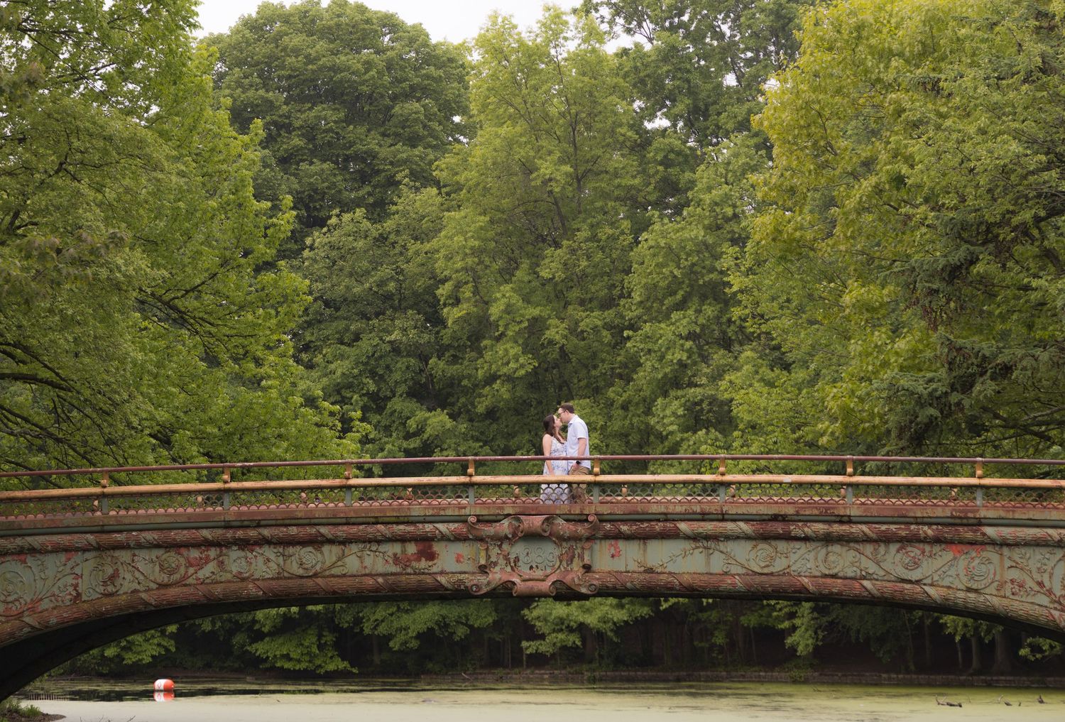 Couple embracing on ornate Bow Bridge in Central Park, surrounded by lush green trees.