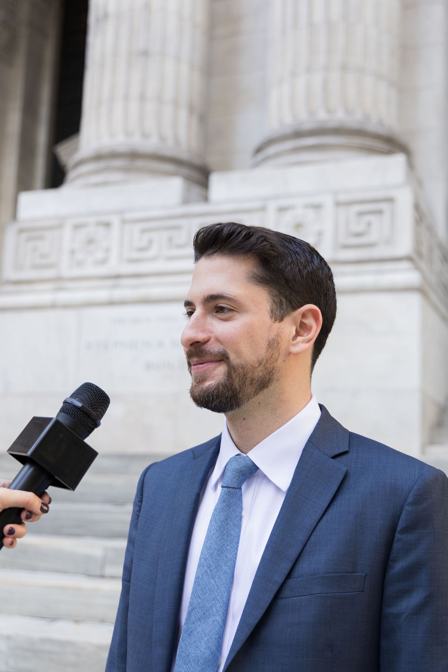 A person in a navy suit and blue tie stands in front of white marble columns at a formal building.