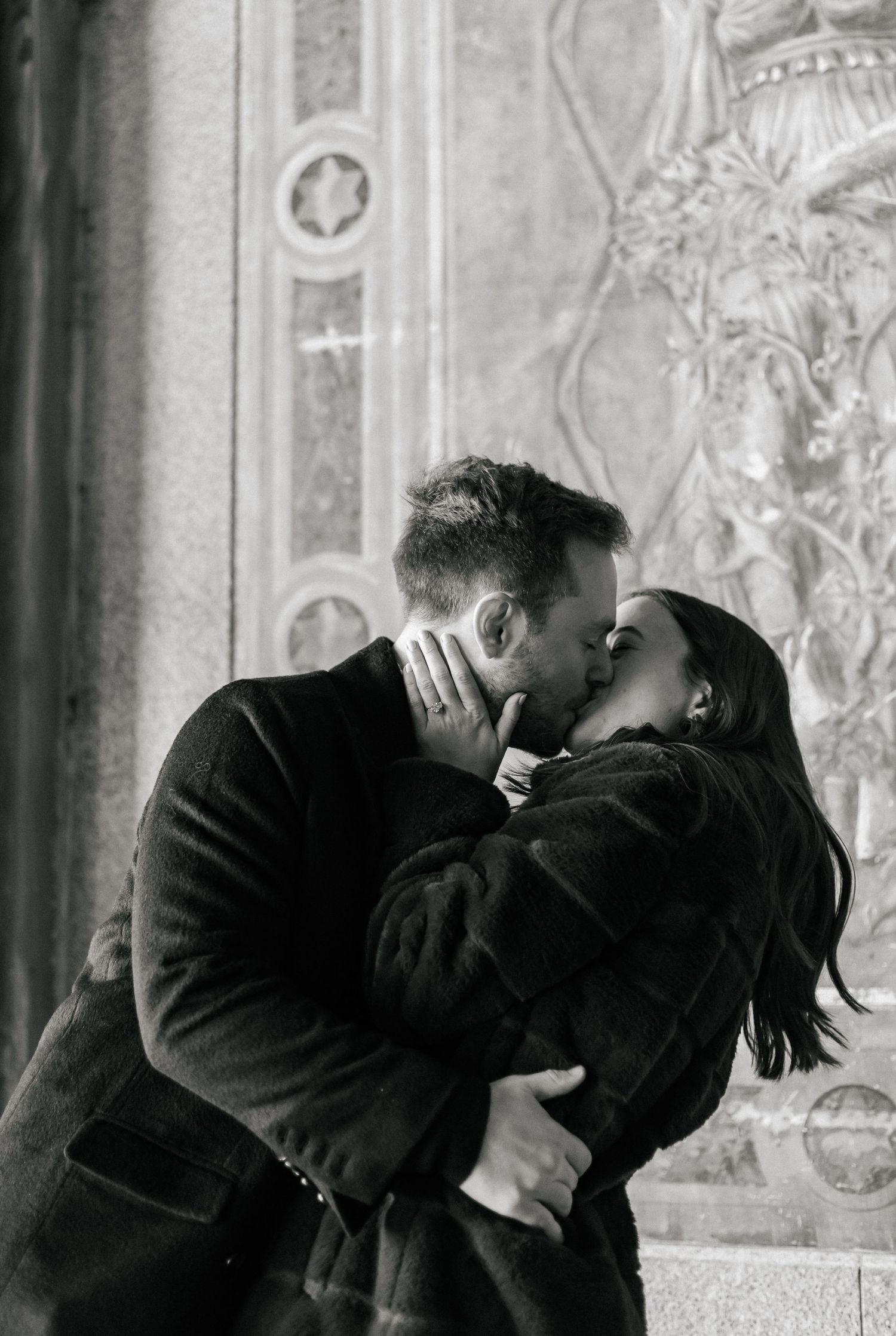 Black and white photo of couple sharing passionate kiss in ornate architectural setting.