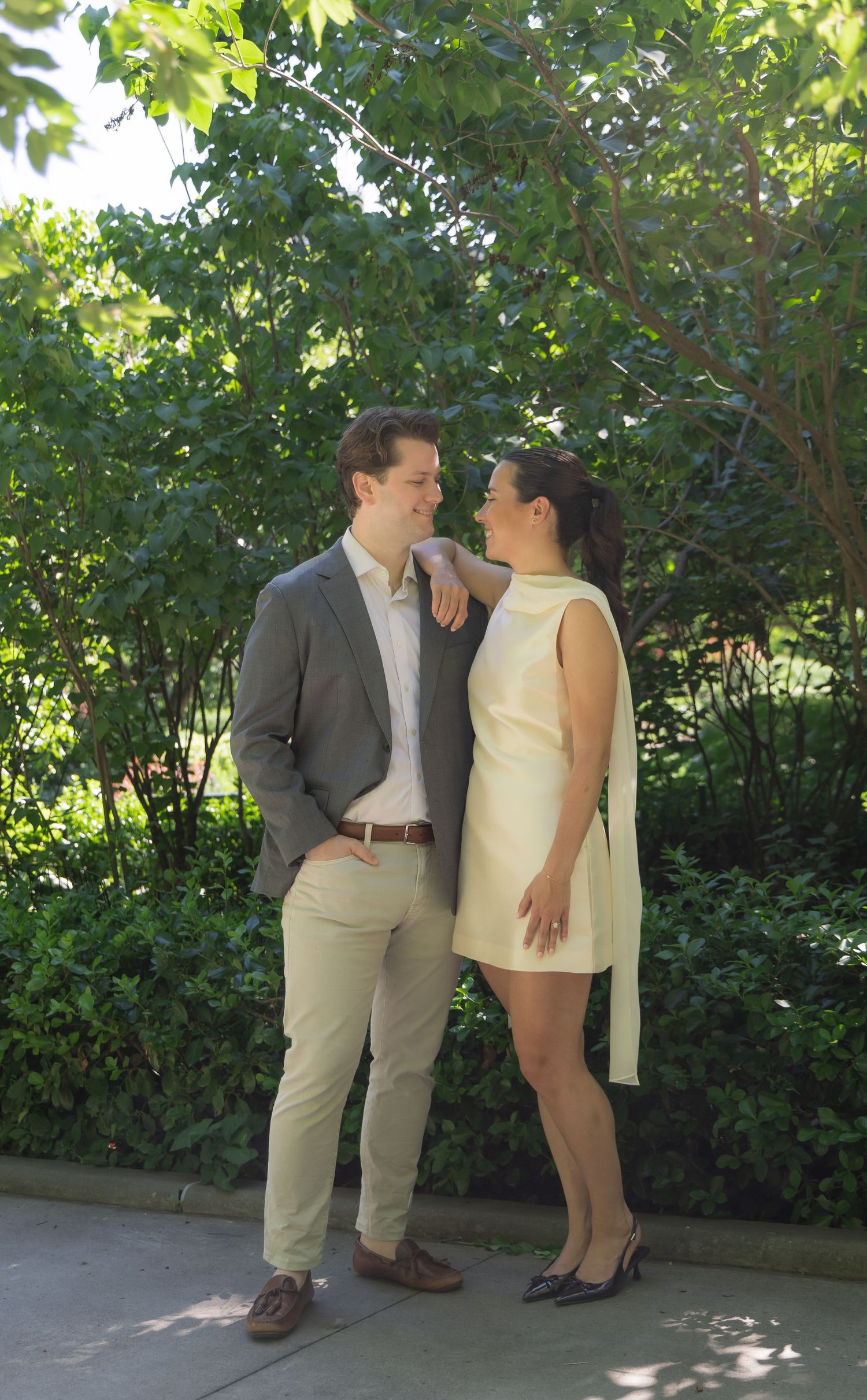 Couple in semi-formal attire standing together outdoors, surrounded by lush green trees and shrubs on a sunny day.