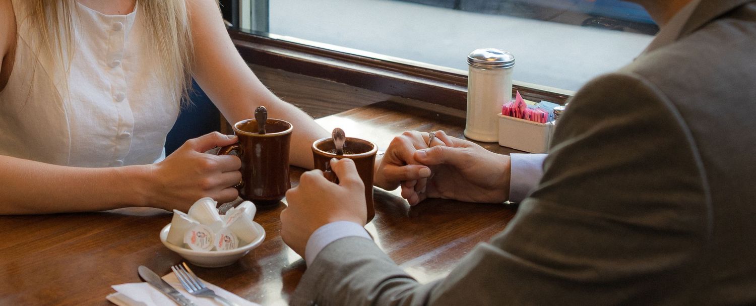 Two people sharing coffee at a wooden cafe table, holding mugs with sugar cubes nearby in a cozy setting.