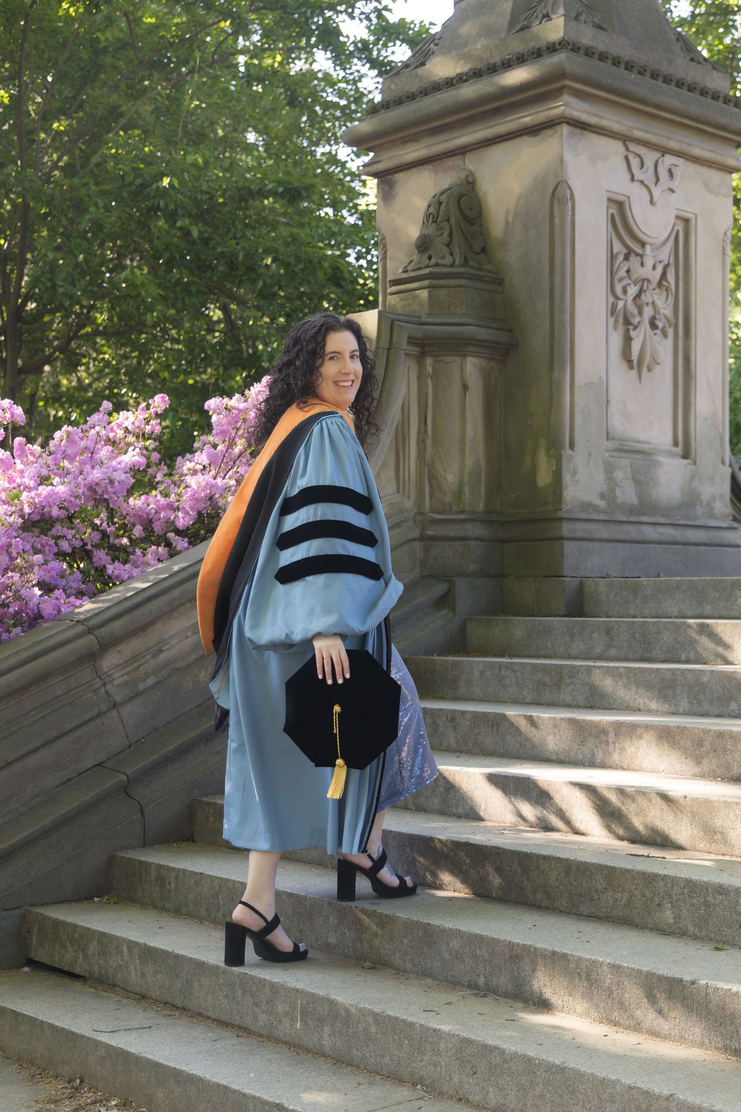 Woman in doctoral graduation gown with orange hood holds cap on stone steps near pink flowers.