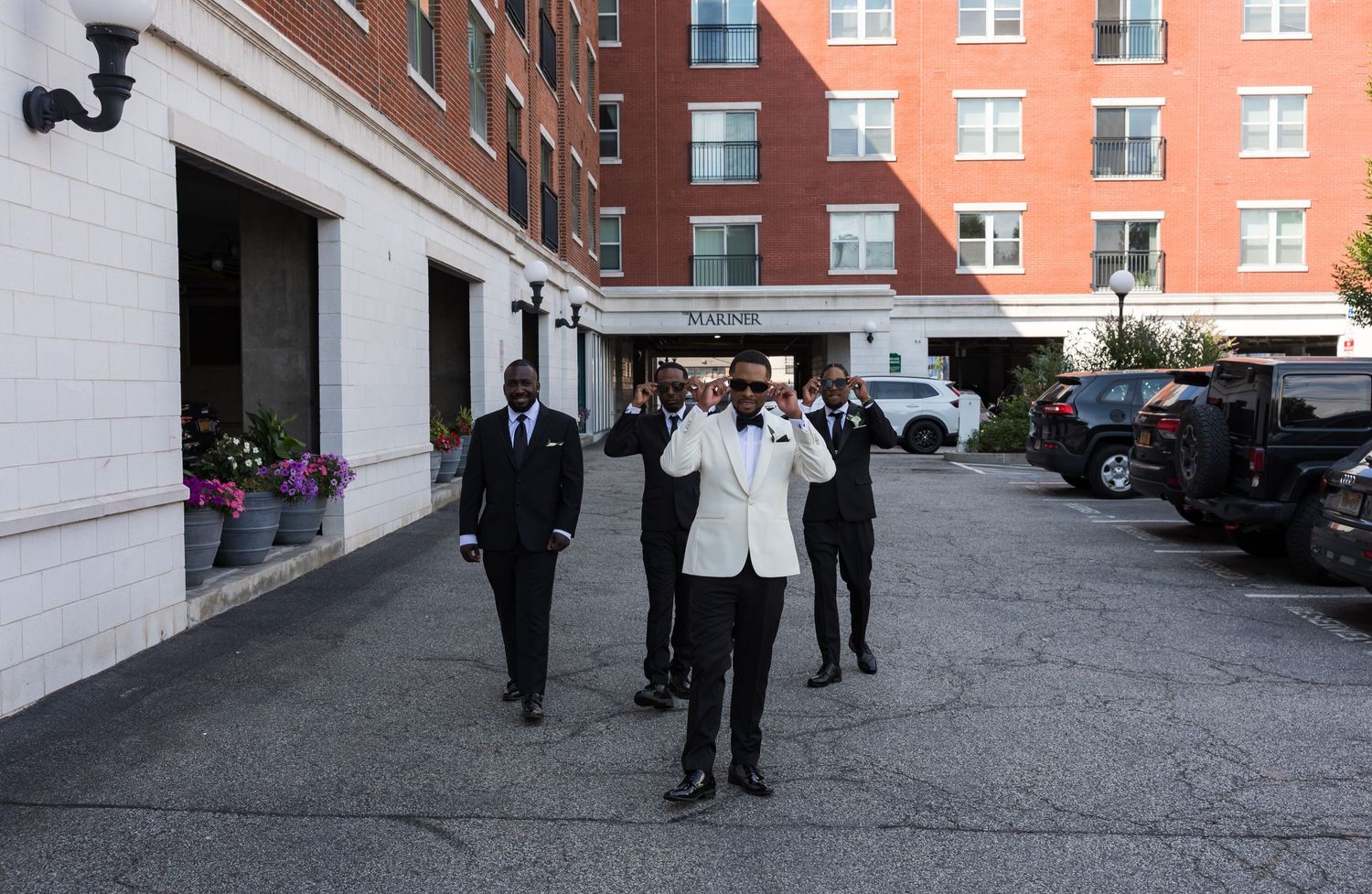 Men in formal attire, including a white tuxedo, walk confidently outside an elegant hotel entrance.