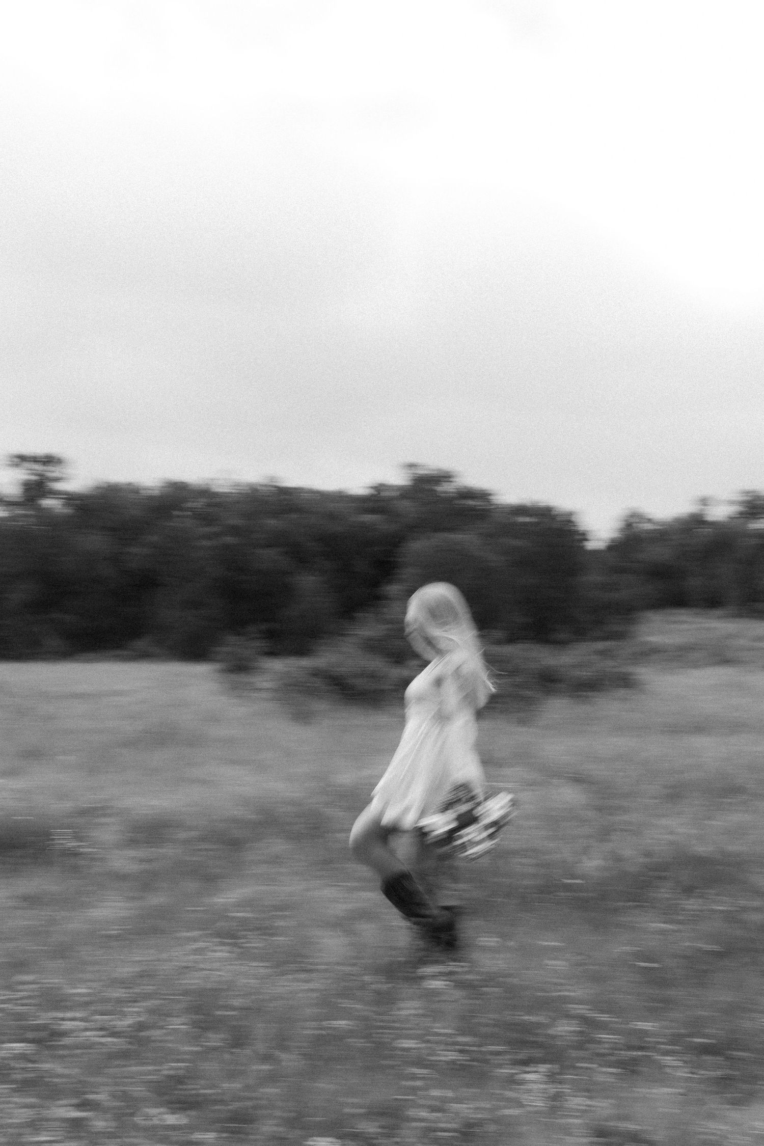 Black and white blurred photo of a woman in a white dress walking through an open field.