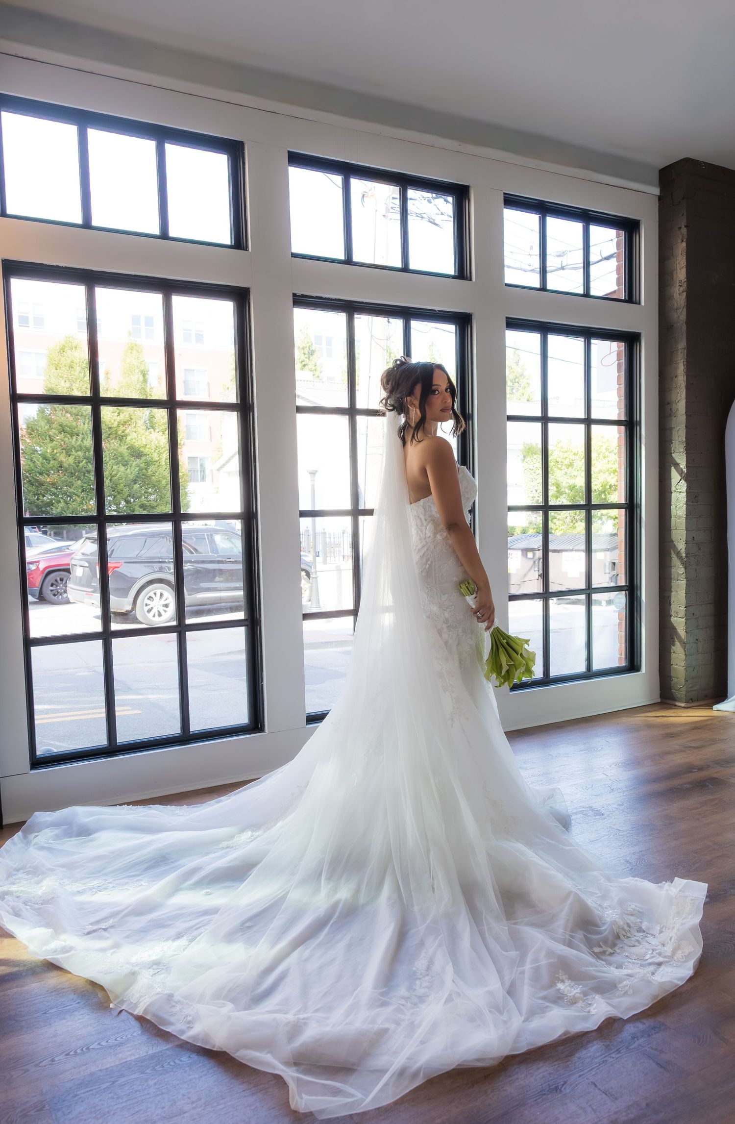 Bride in a flowing white wedding gown with long train stands by large windows holding a bouquet.