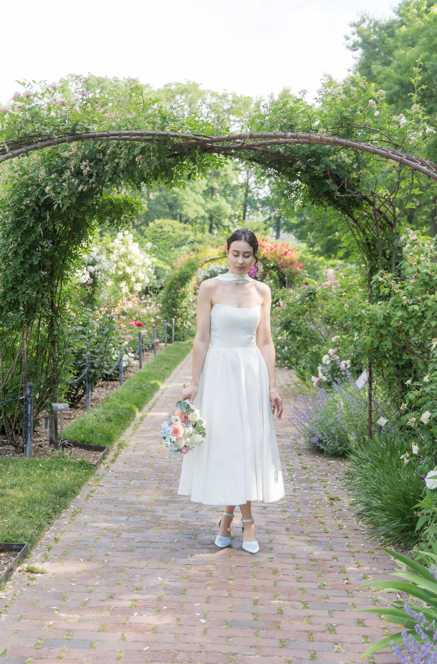 Bride in white strapless midi dress holding bouquet under floral garden arch on brick pathway.