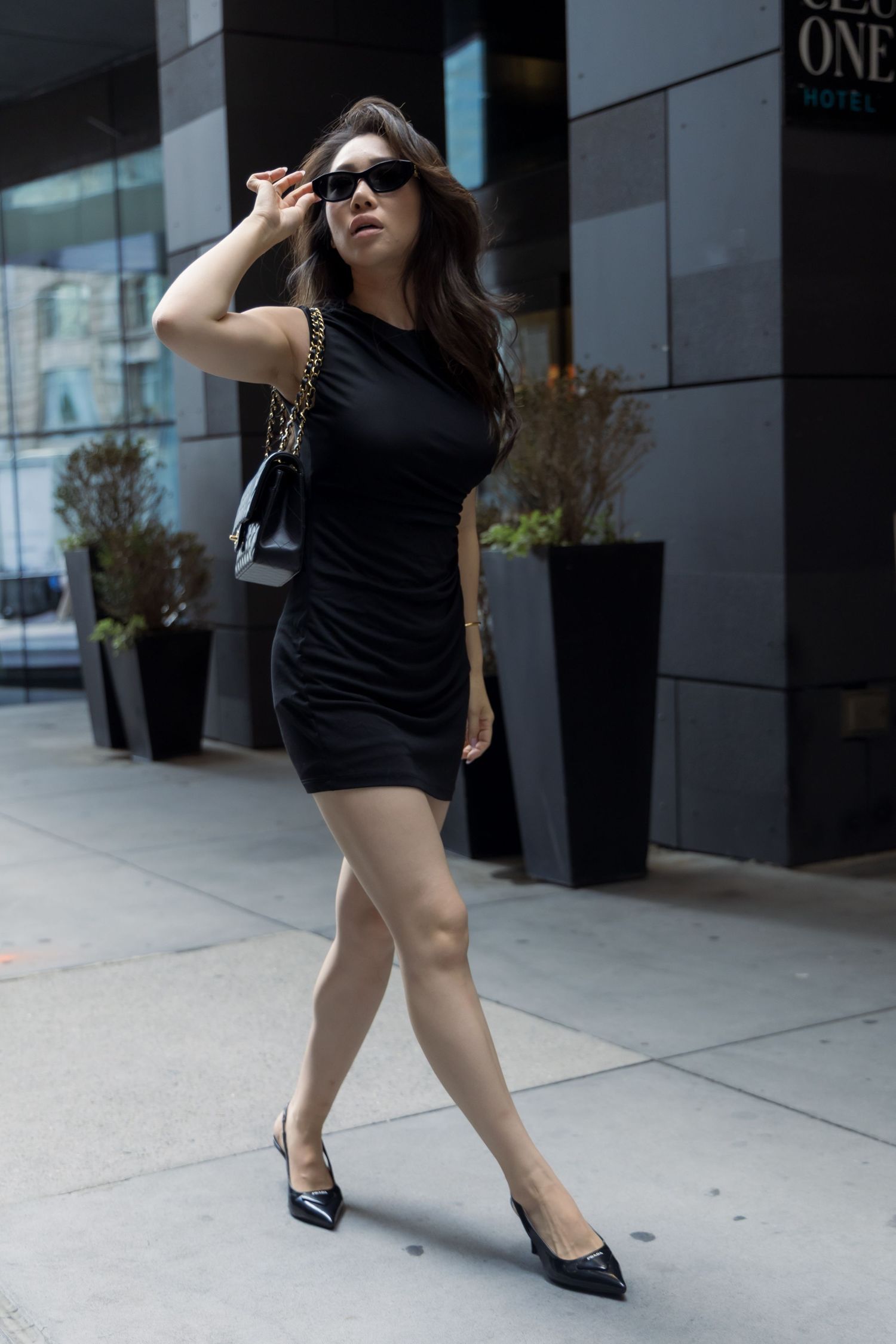 Stylish woman in black mini dress and sunglasses poses on urban sidewalk with tall planters in background.