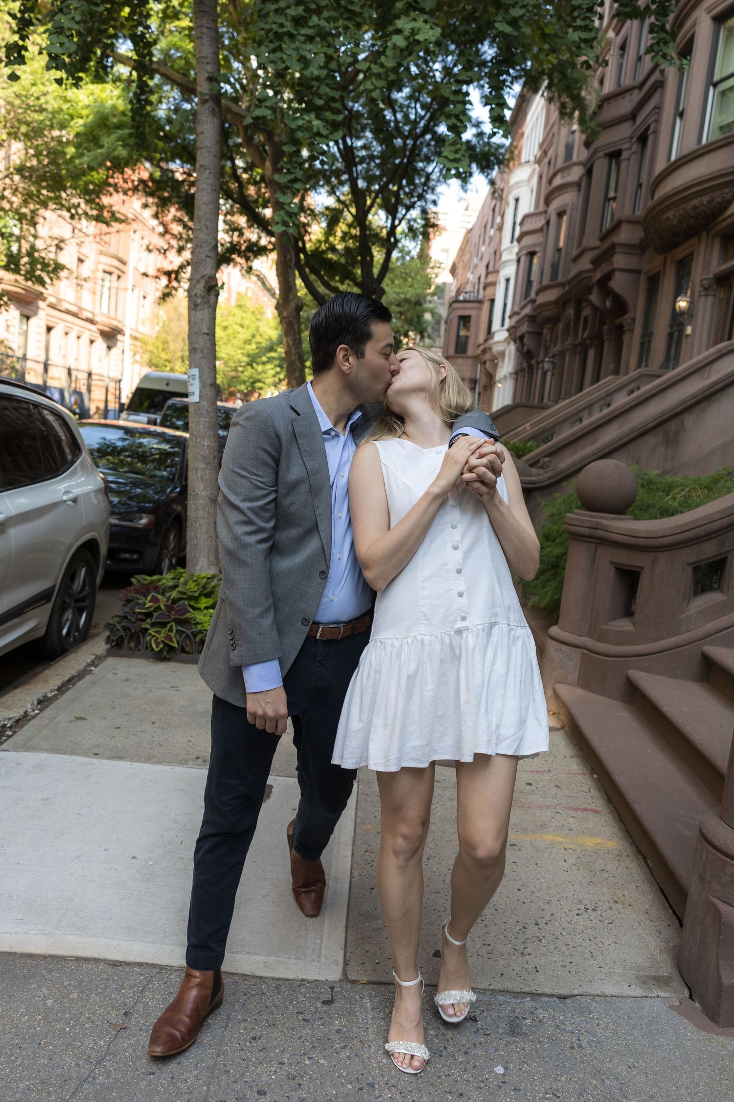 Couple sharing a romantic kiss on a sunny urban sidewalk, woman in white dress holding hands with man in gray blazer.