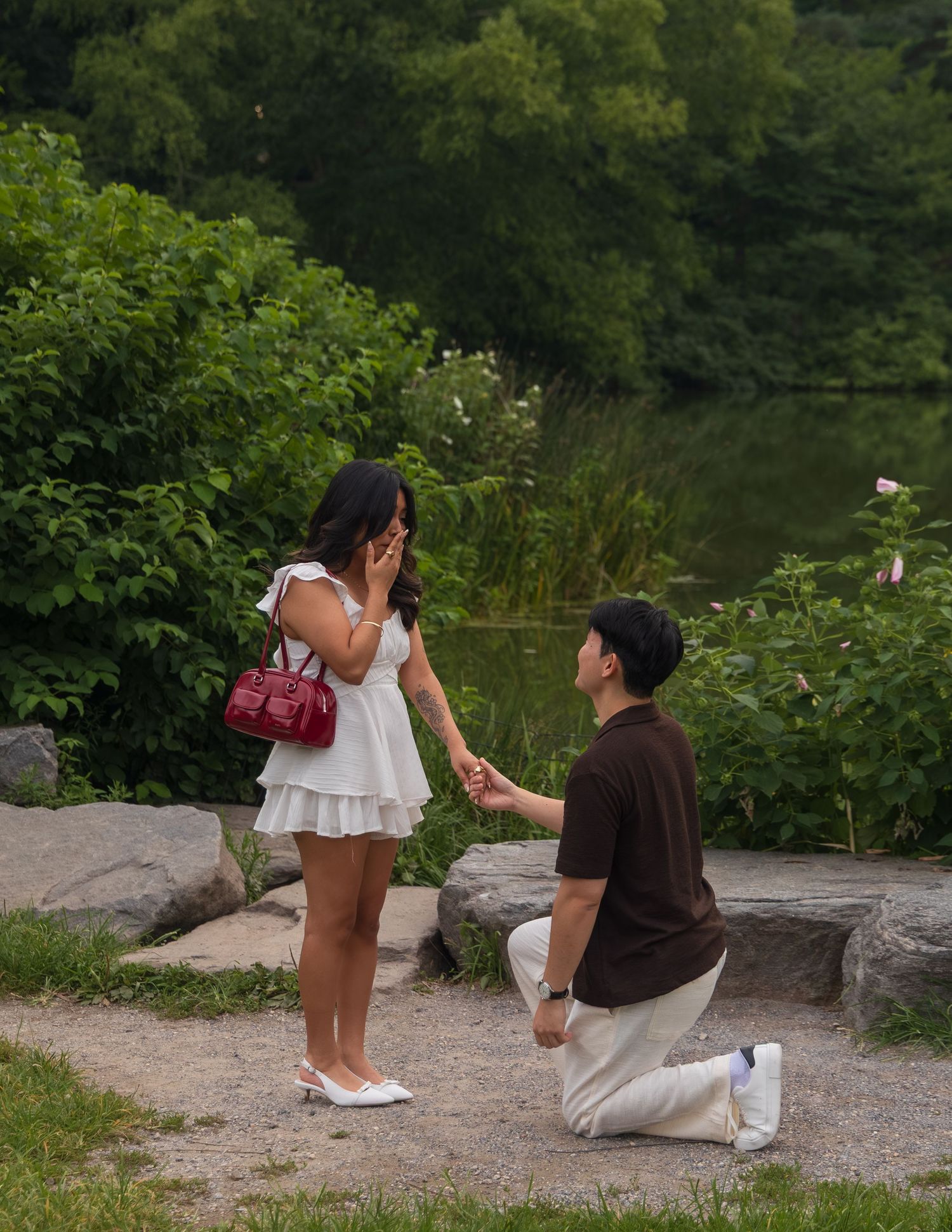 Man kneeling on one knee proposing to woman in white dress by a lake in a lush green park.