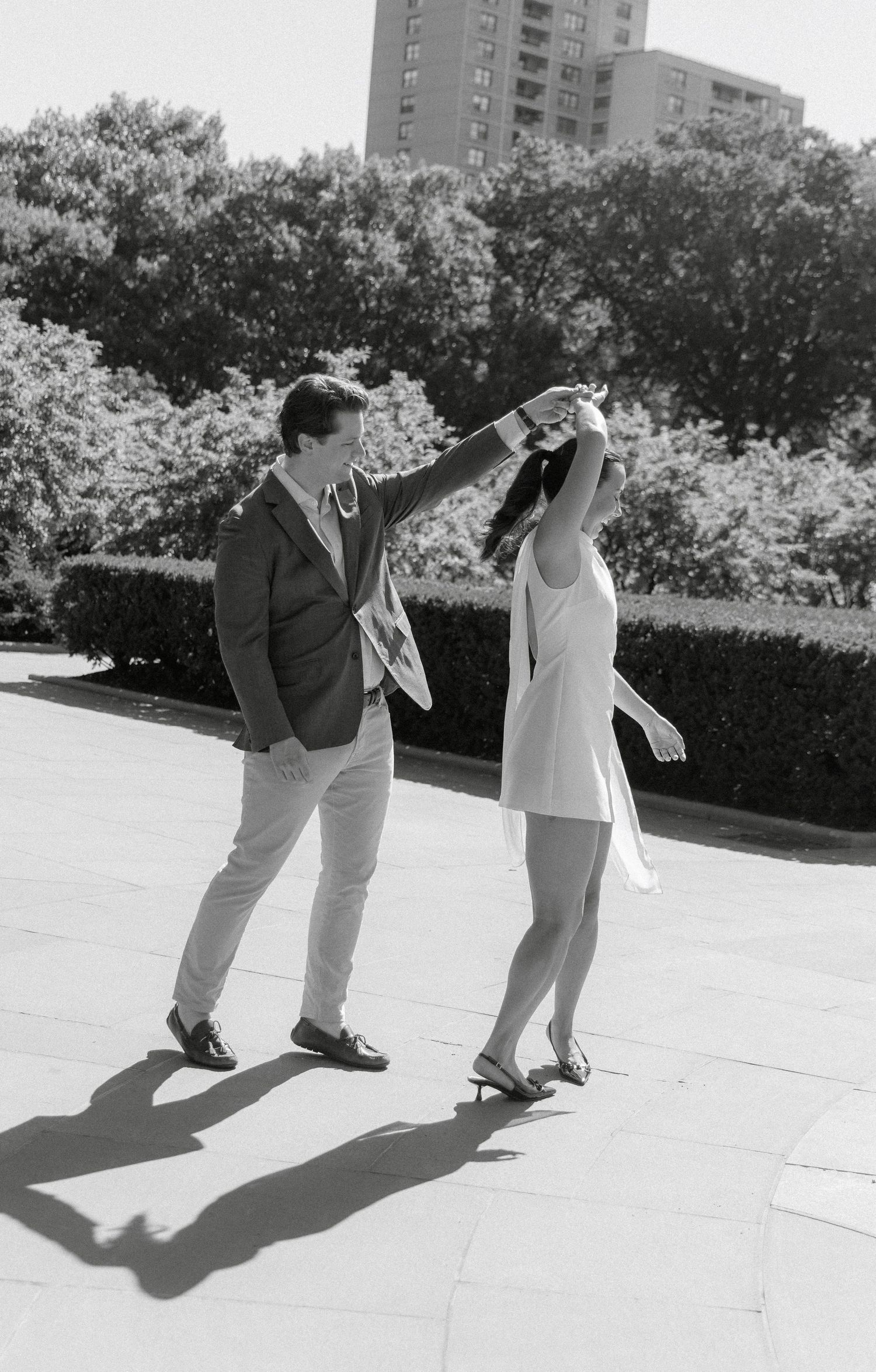 Black and white photo of a couple dancing outdoors, man spinning woman in a white dress, trees and building in background.