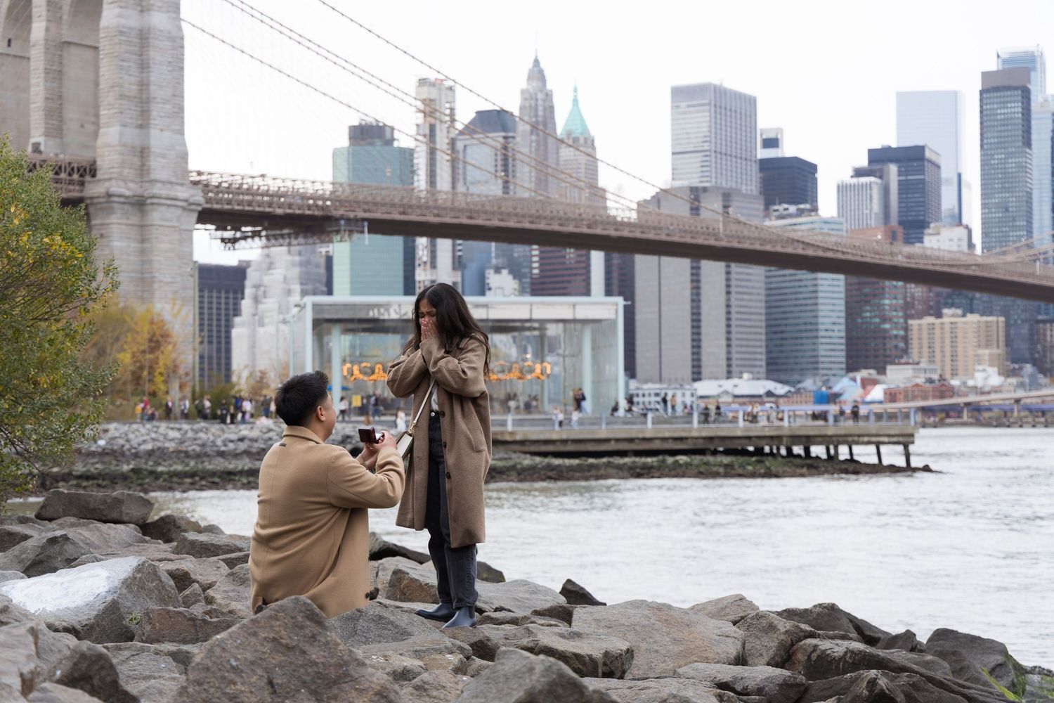 A romantic proposal scene with the Brooklyn Bridge and Manhattan skyline in the background.