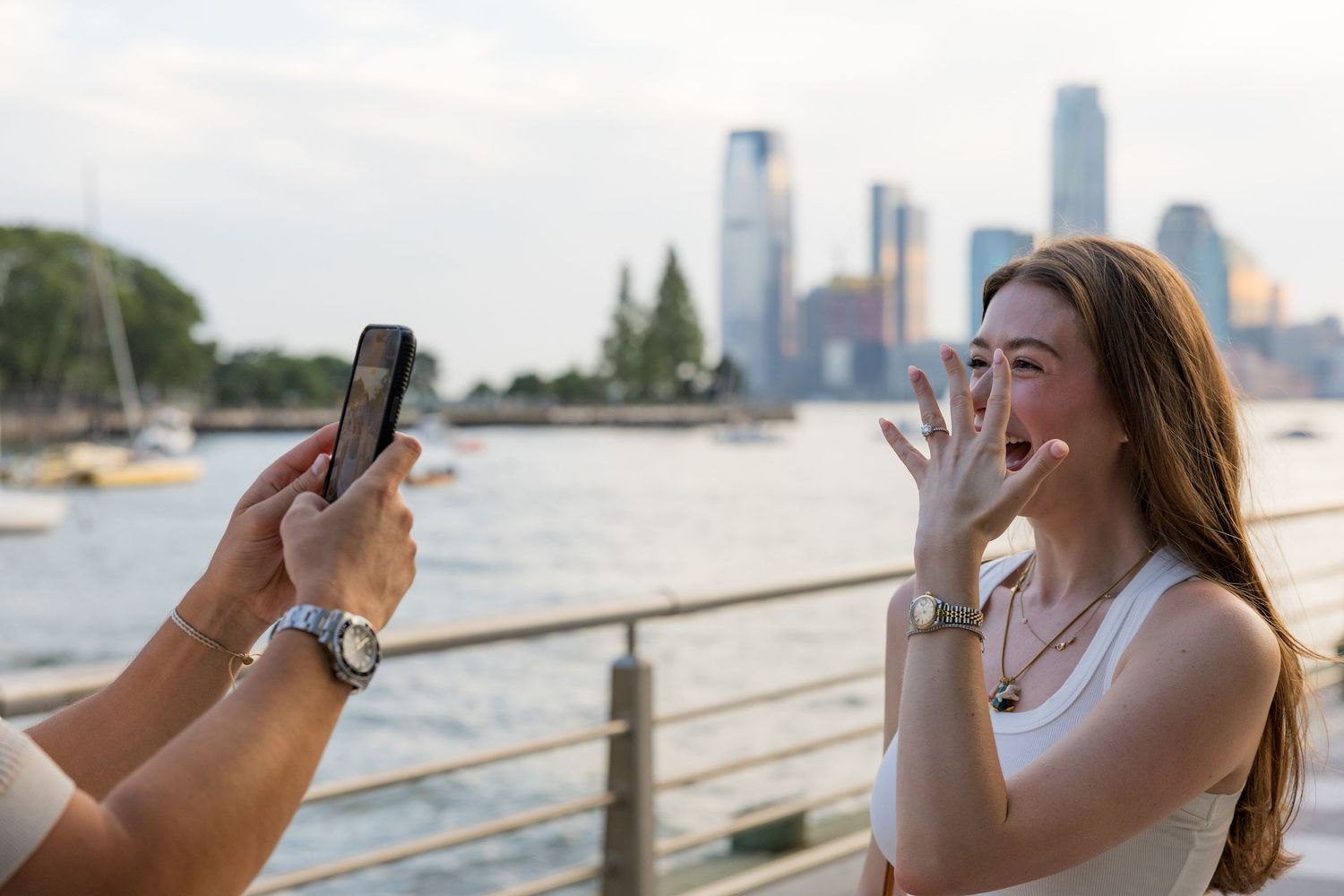 Woman laughing as friend photographs her with smartphone by waterfront with city skyline in background.