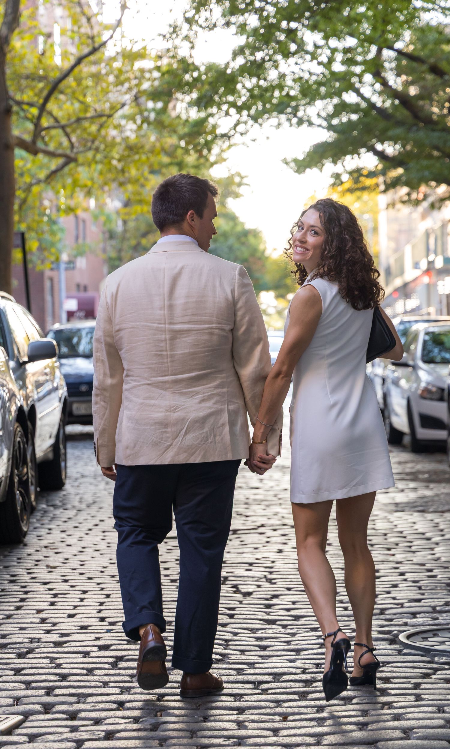 A couple holds hands while walking on cobblestone road in the midst of brownstones on Bank Street in the West Village.