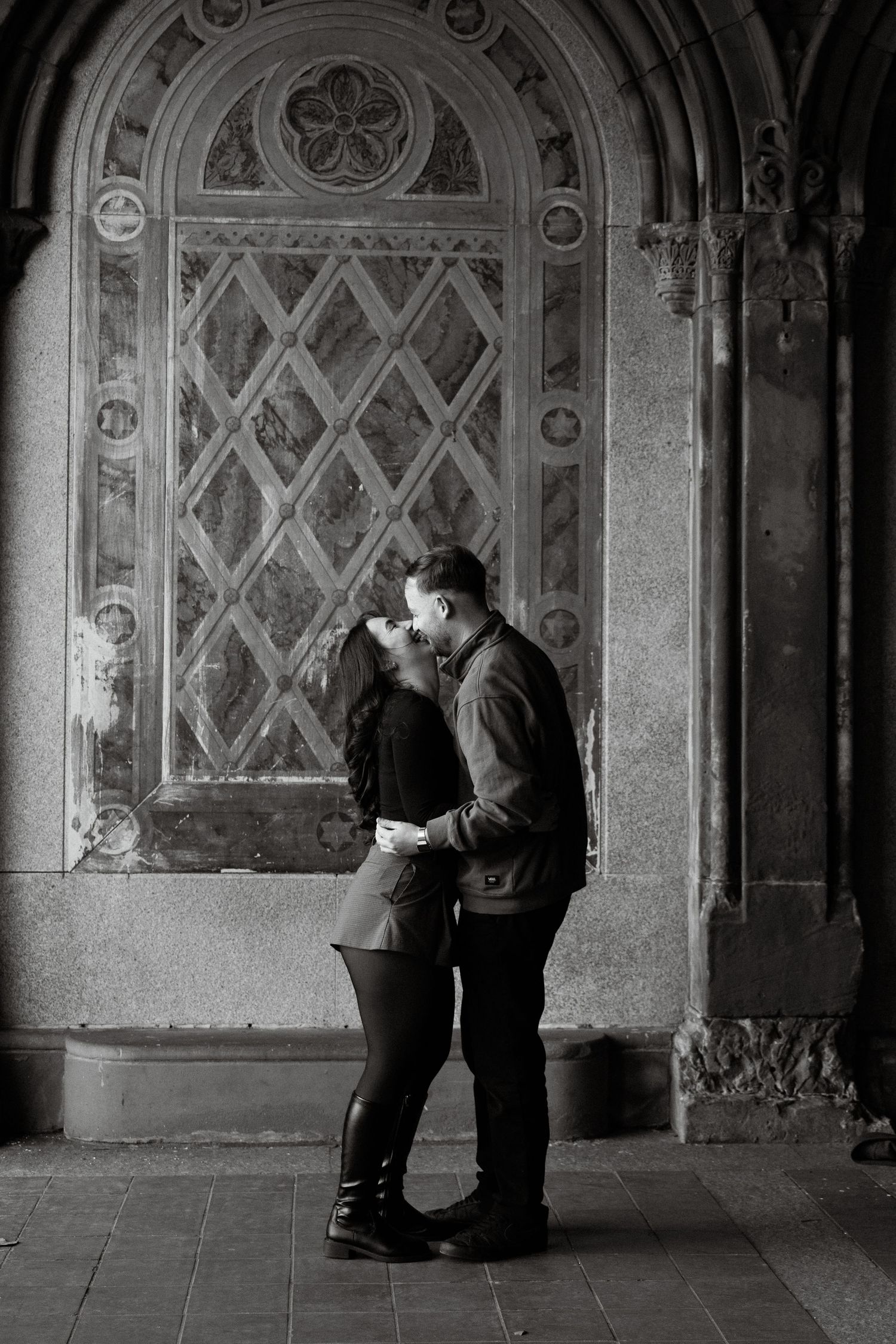A romantic couple shares an intimate moment in black and white beneath an ornate archway at Bethesda Terrace