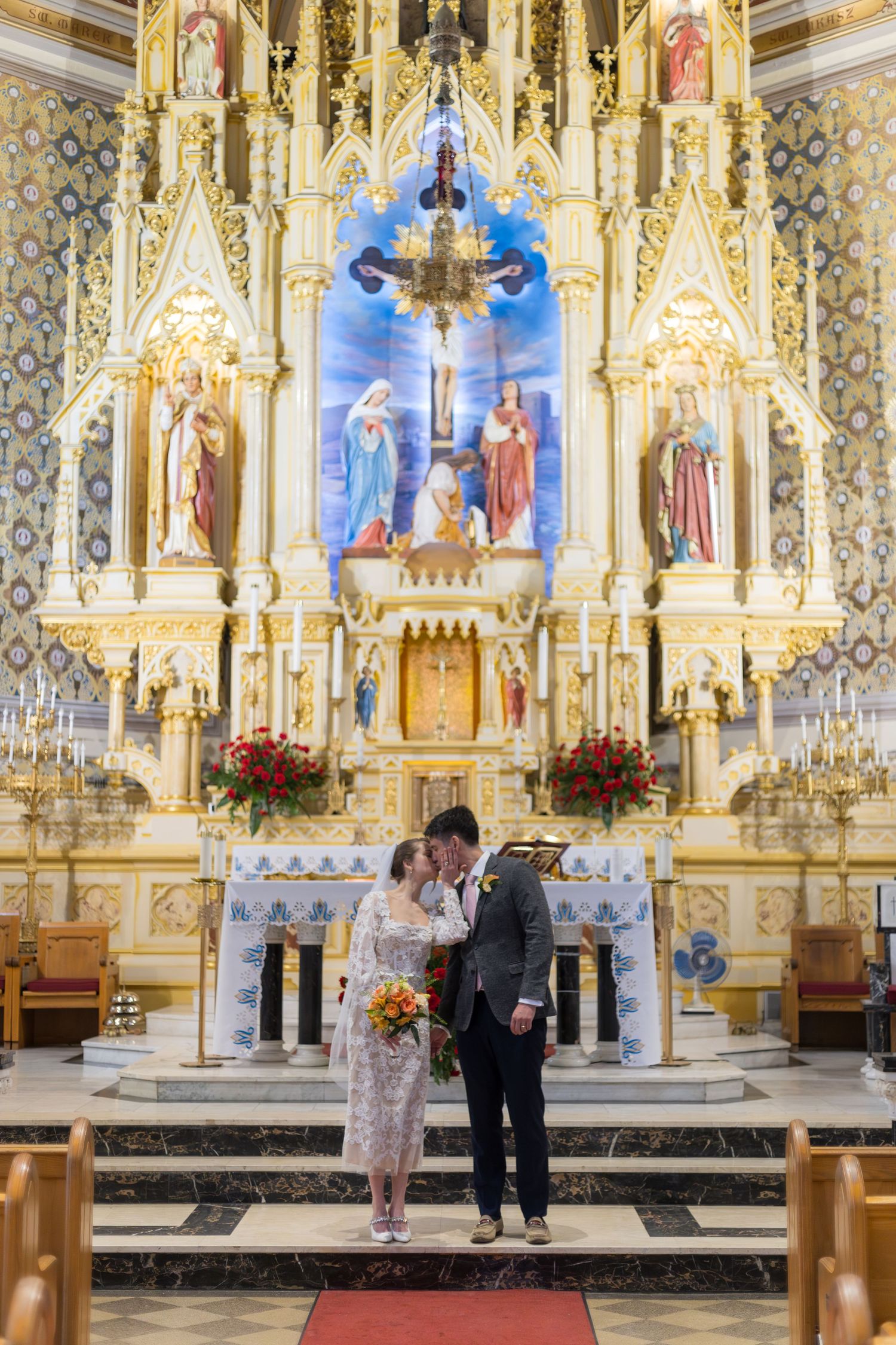 Couple shares a kiss at ornate Catholic church altar adorned with gold details and religious statues on their wedding day.