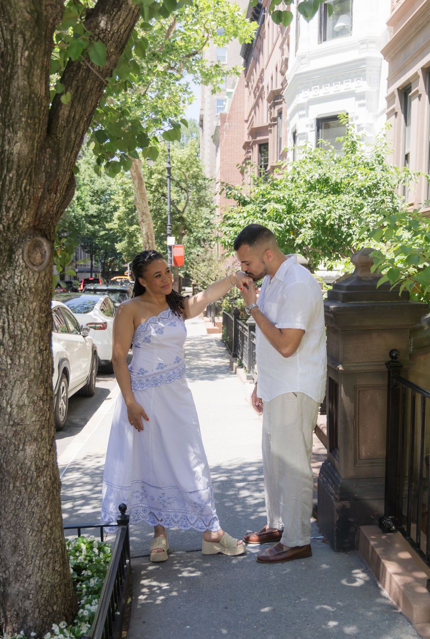 A man kisses a woman's hand on a sunny tree-lined sidewalk, both dressed in white, in an urban neighborhood.