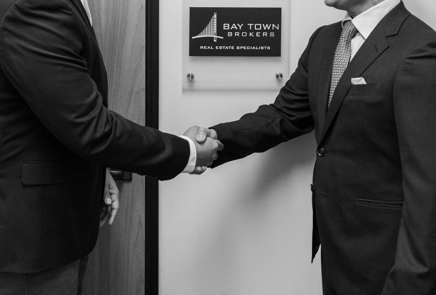 Two people in business suits shake hands in front of an office wall with a company sign.