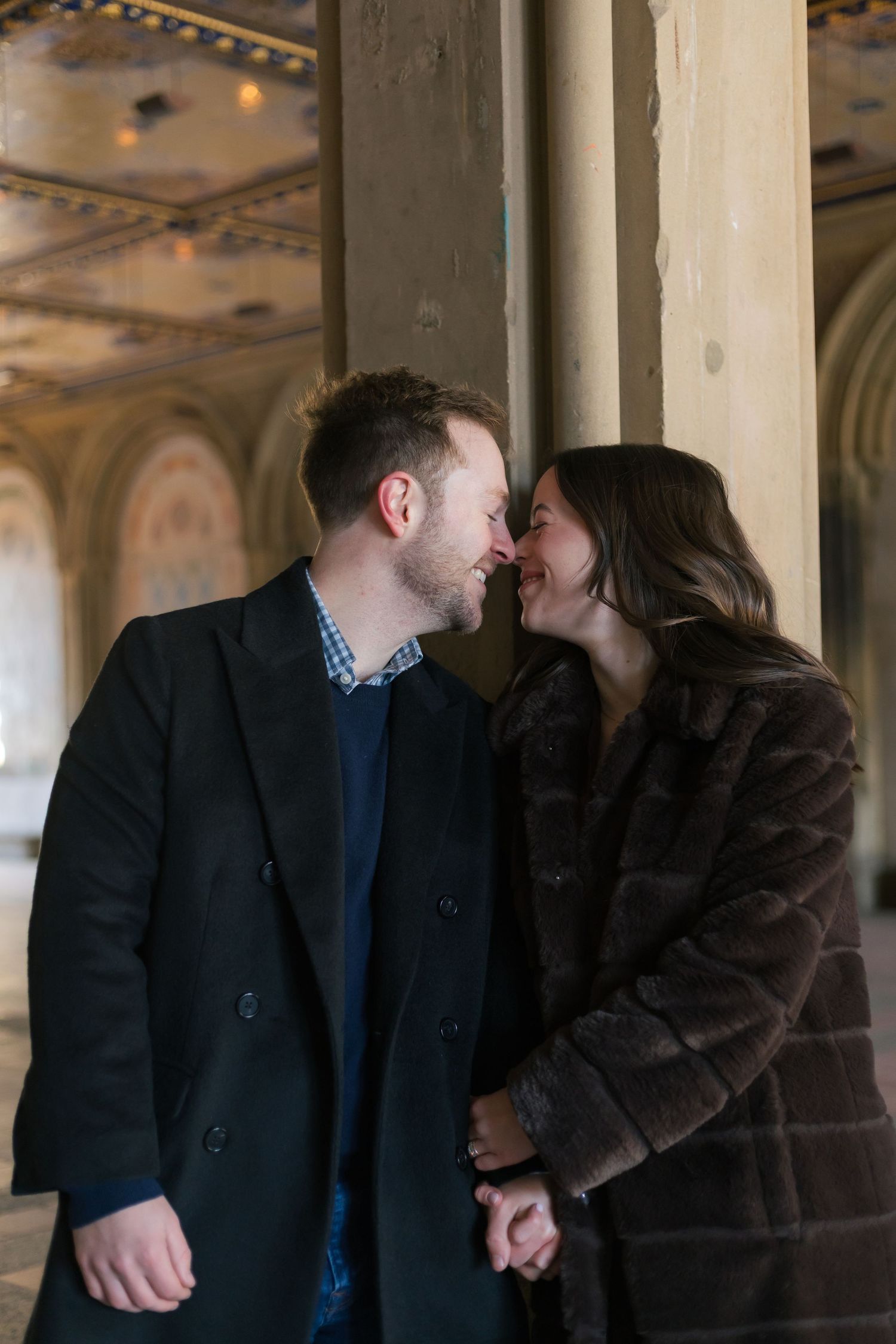 Couple sharing a romantic moment, nearly kissing, dressed in winter coats near a stone column indoors.