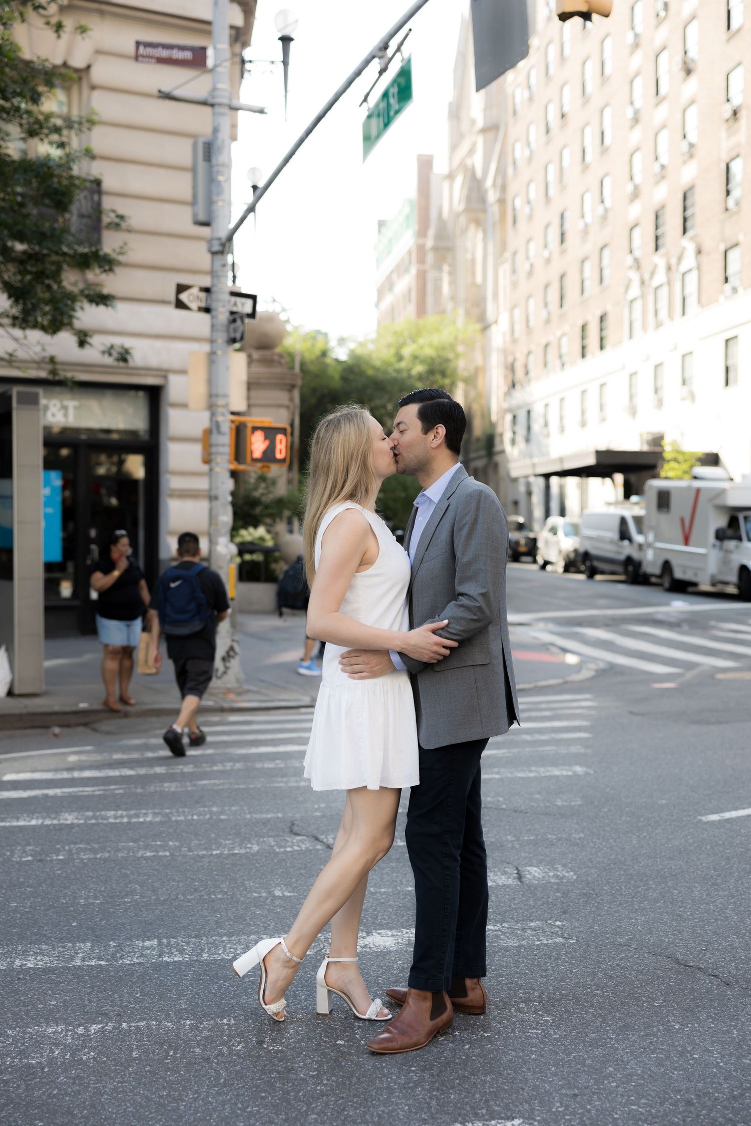 Couple sharing a romantic kiss on a busy New York City crosswalk, woman in white dress and man in gray blazer.