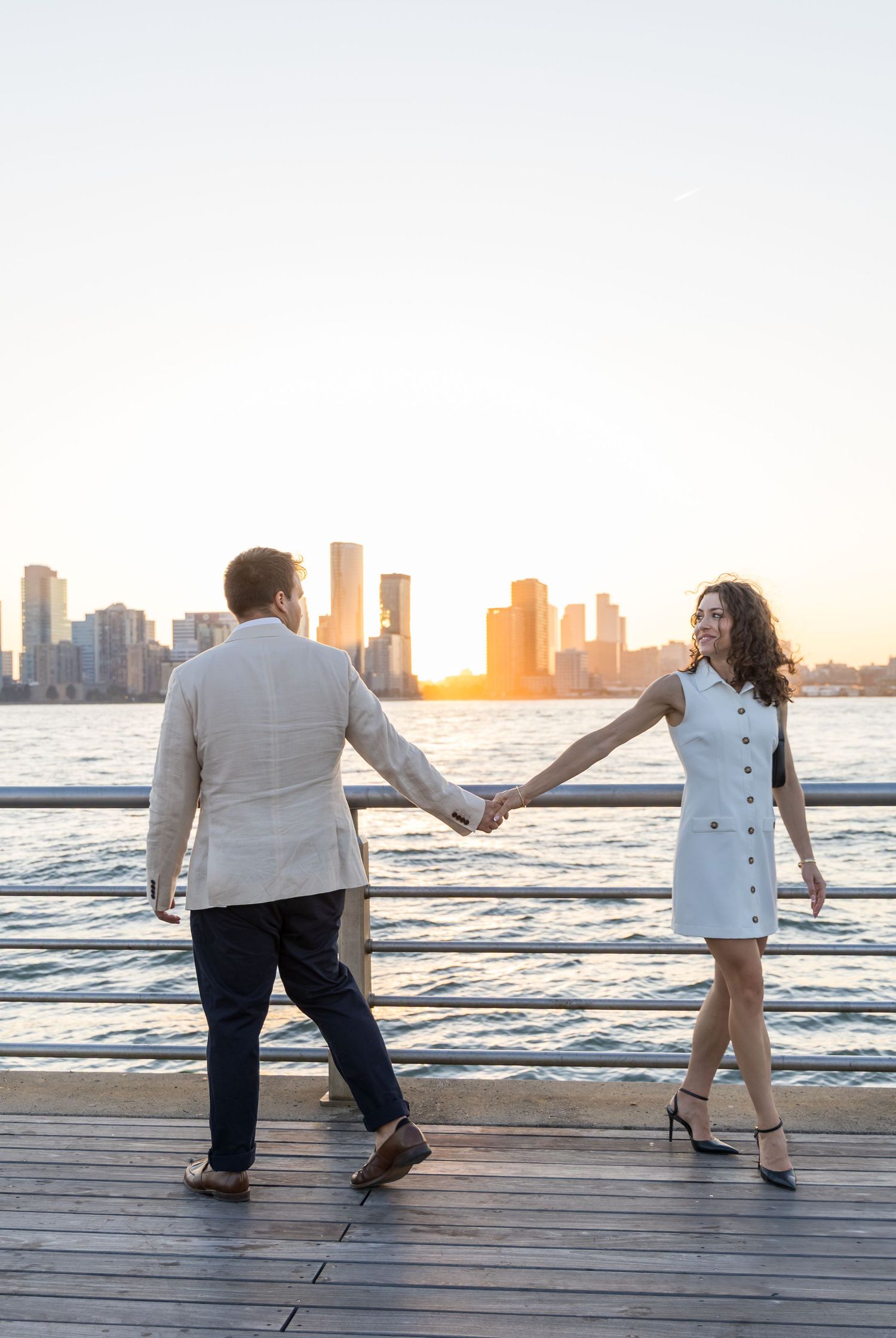 A couple playfully poses on a wooden boardwalk by the water with a city skyline at sunset.