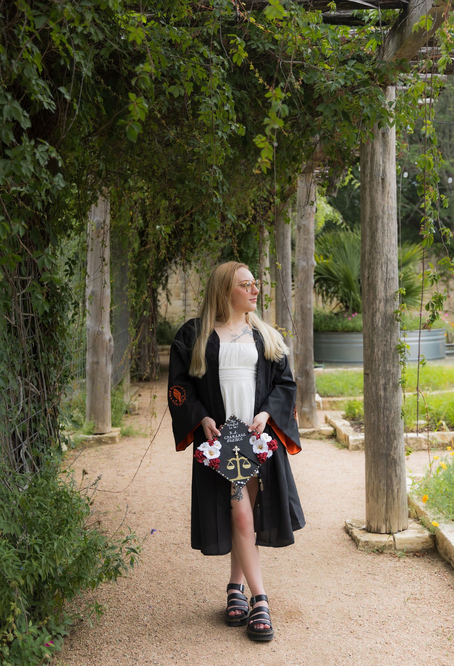 Blonde graduate in black gown walking under green pergola holding decorated graduation cap.