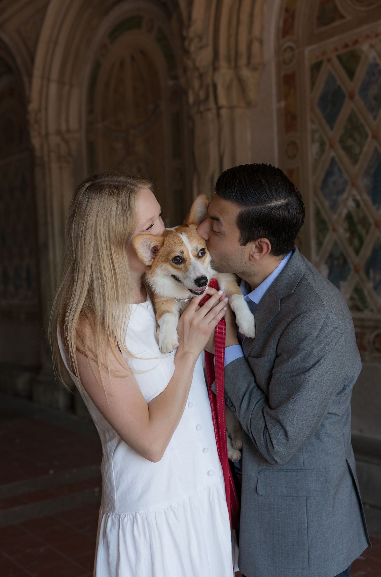 Couple in white dress and gray suit kissing a Corgi dog inside an ornate stone archway.