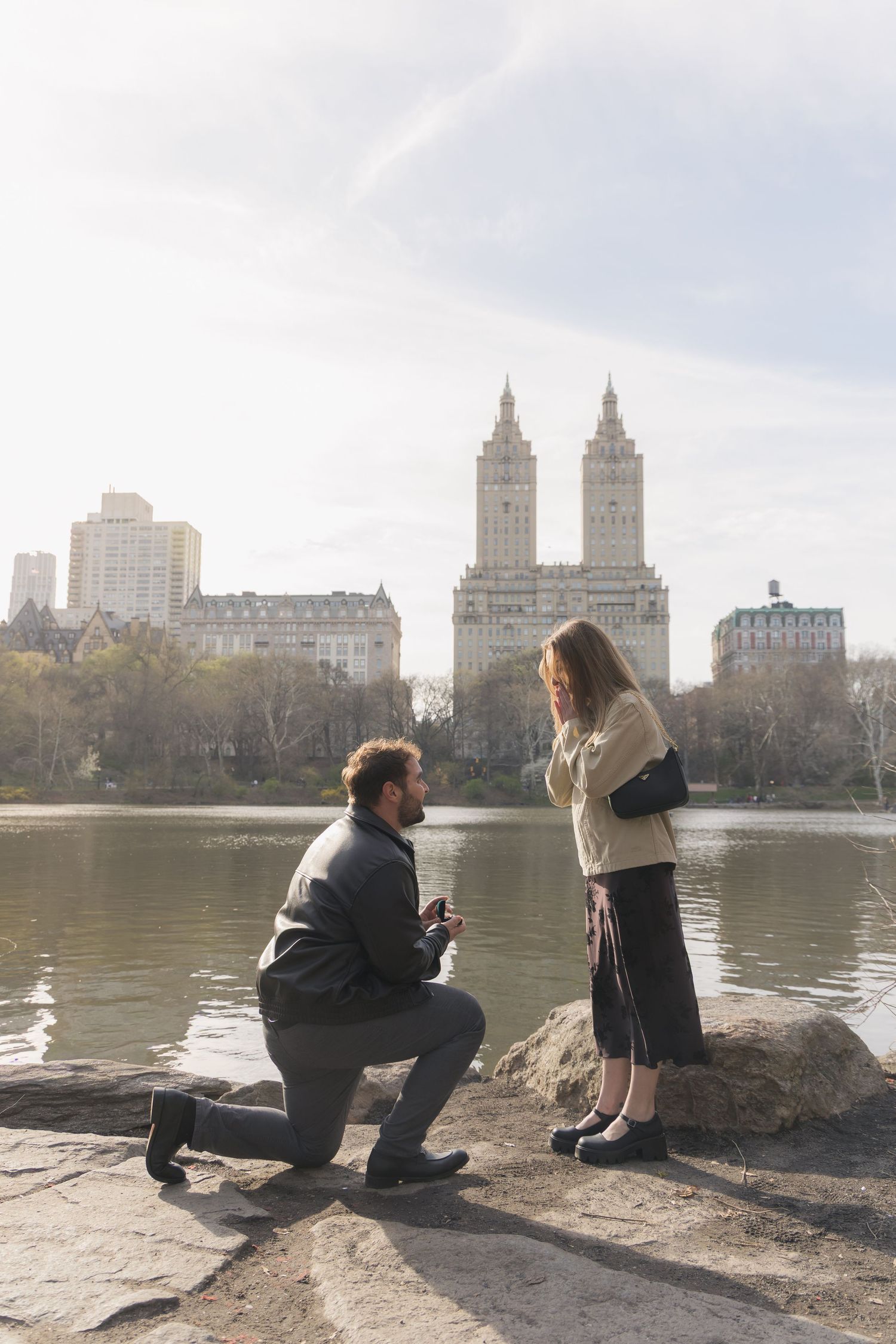 Man proposes on one knee to woman by Central Park lake with iconic San Remo building in NYC background.