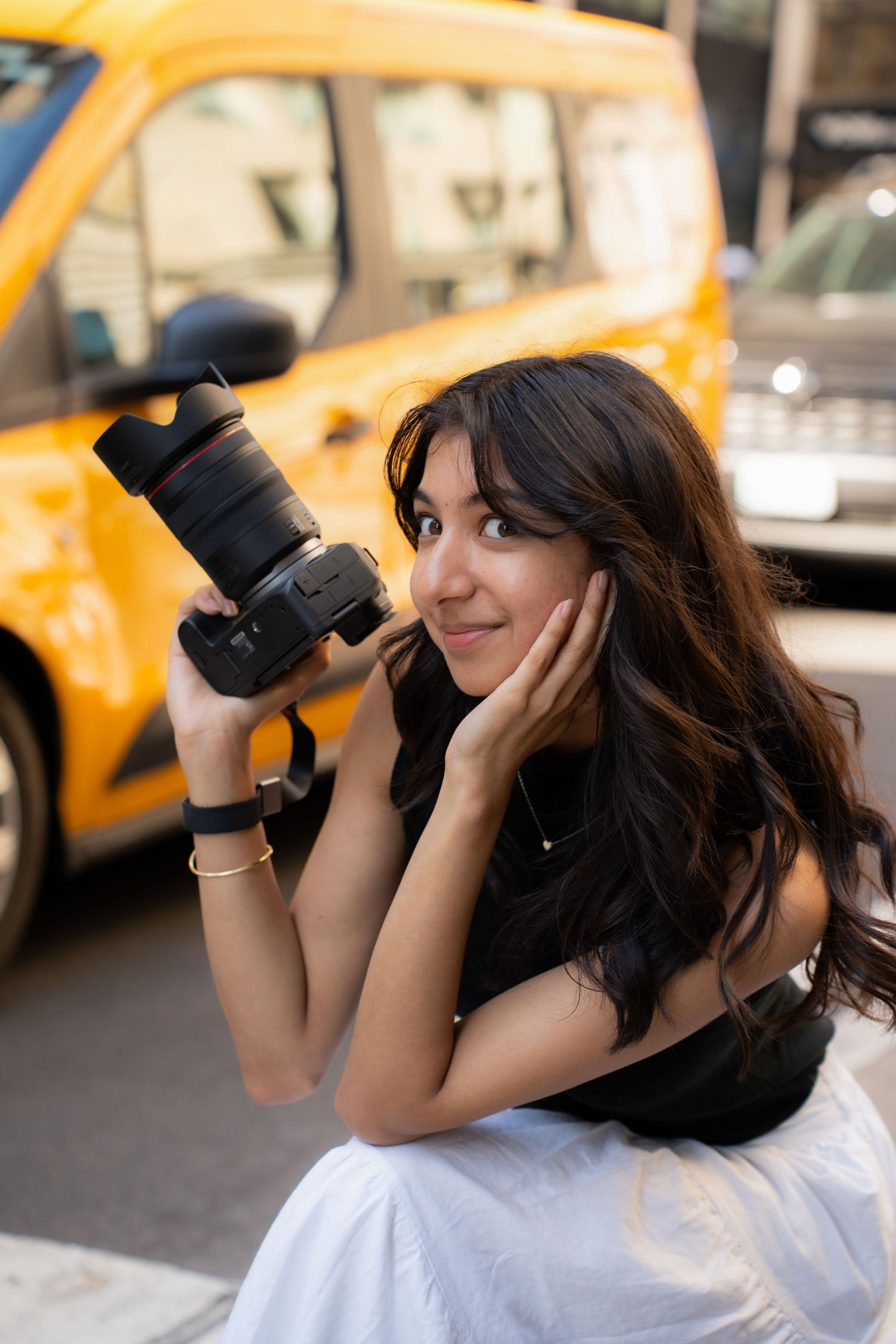 A photographer in a white top uses a professional camera to take photos on a city street with yellow taxis in the background.