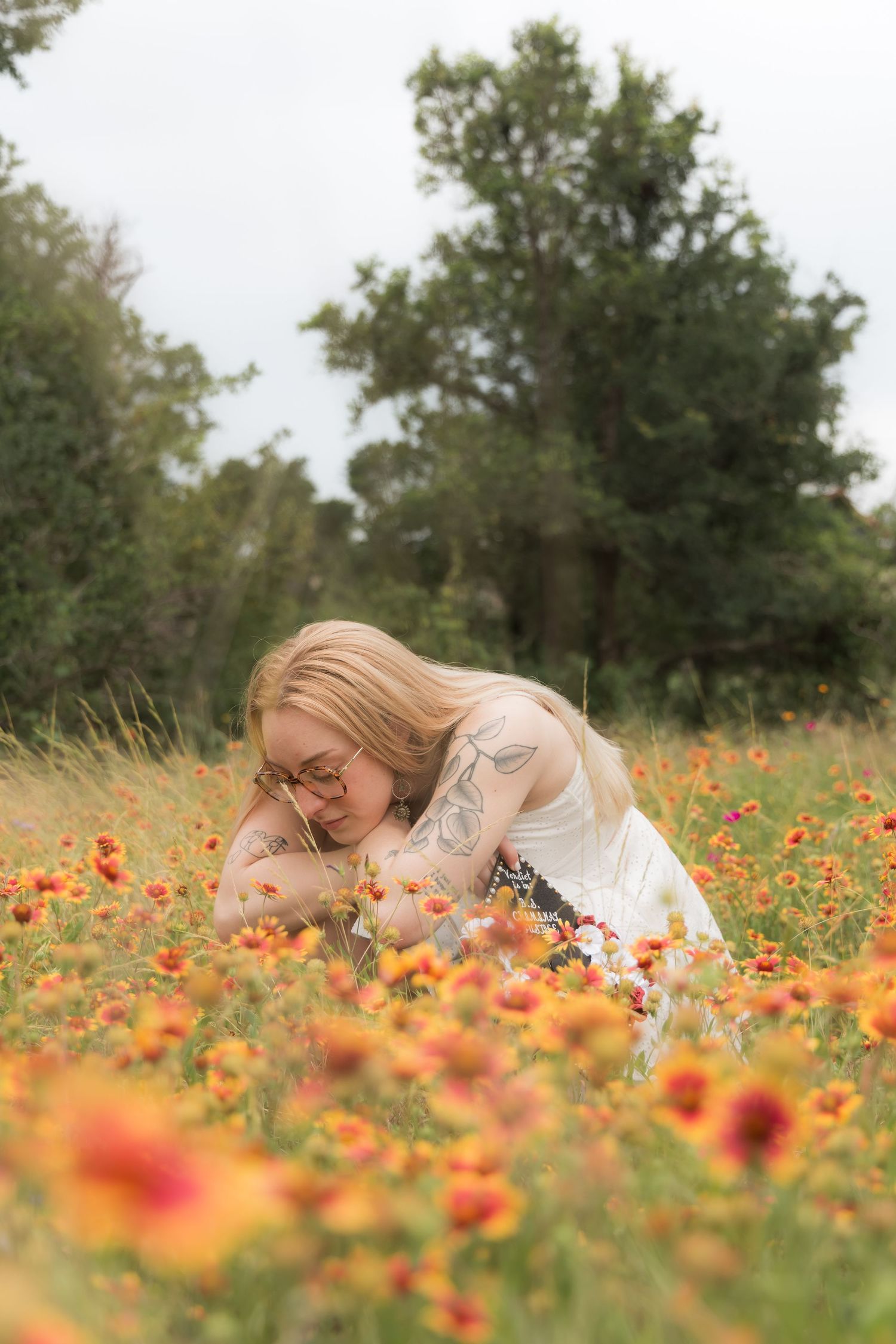 Blonde woman with tattoos lying in vibrant orange wildflower field smelling flowers.