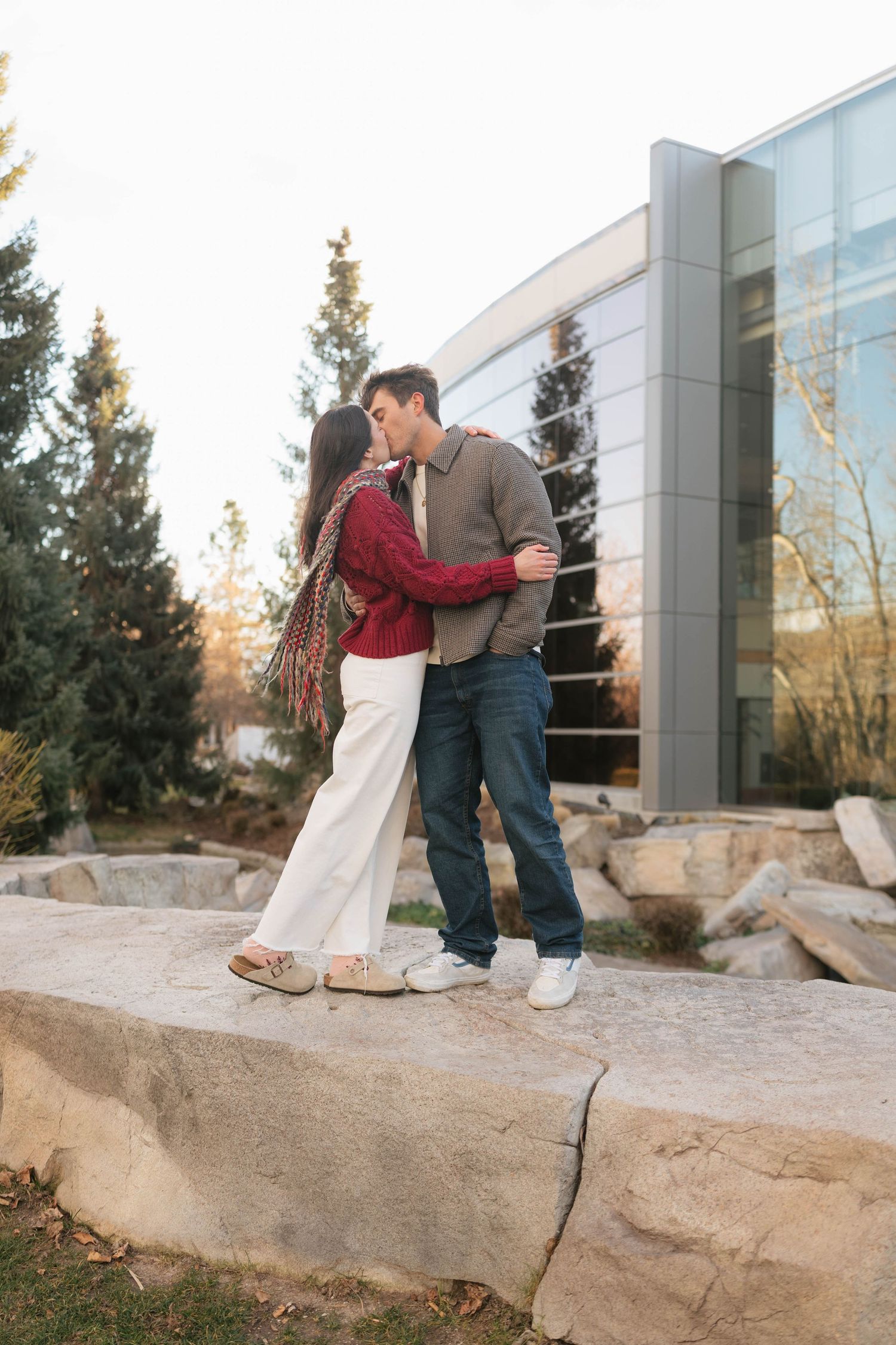 A couple shares an intimate moment together while sitting on a large rock outside a modern building at sunset.