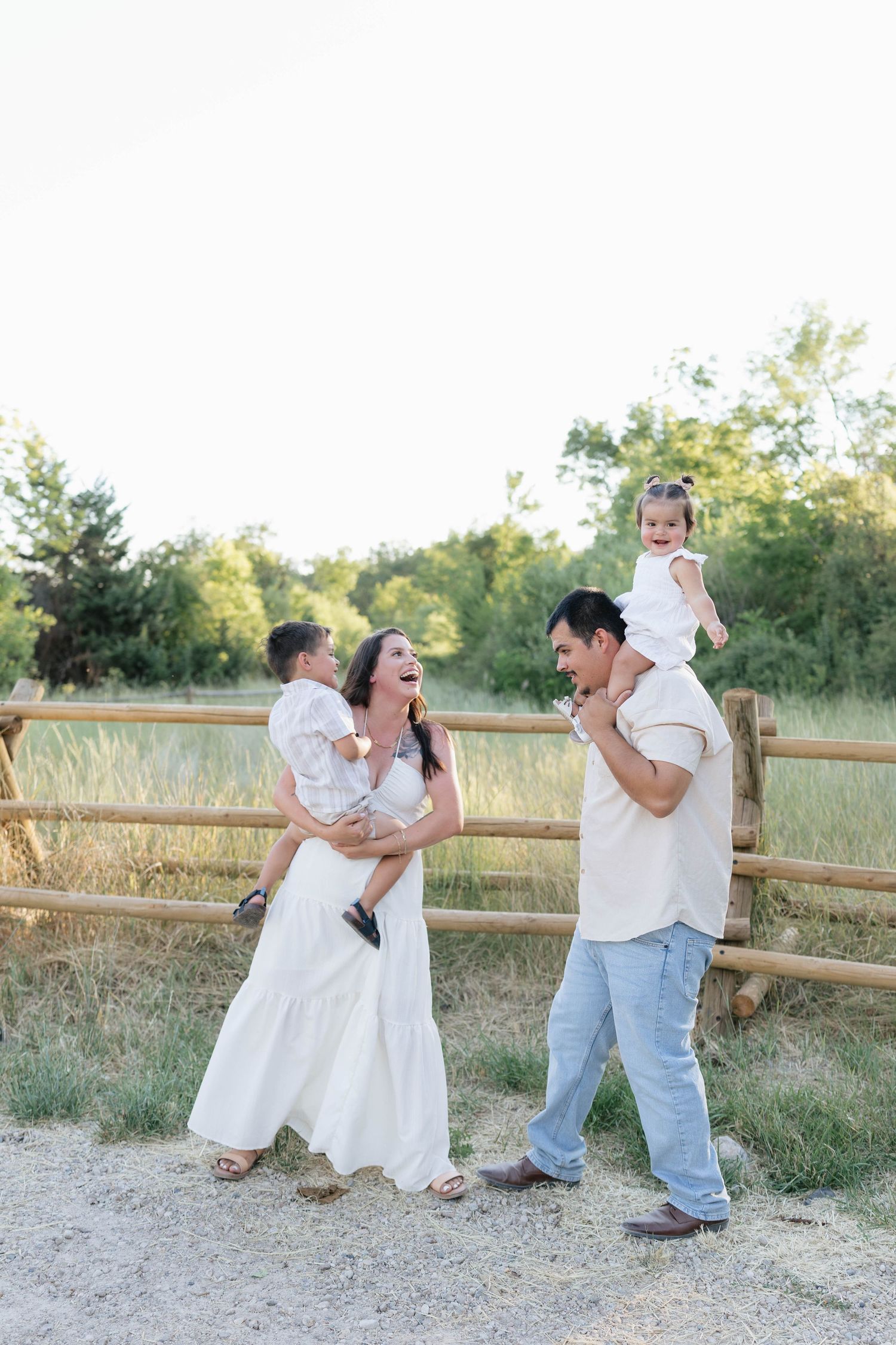 Family of four laughing outdoors near a wooden fence, parents holding young children, dressed in white and light blue.