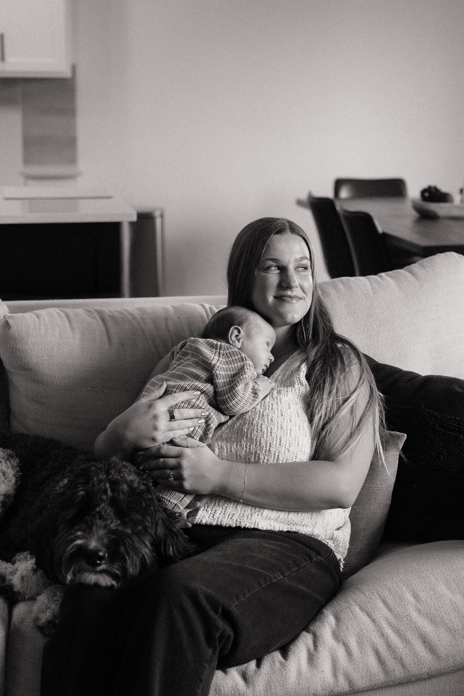 Black and white photo of a young woman sitting on a sofa, cradling a newborn baby with a fluffy dog beside her.