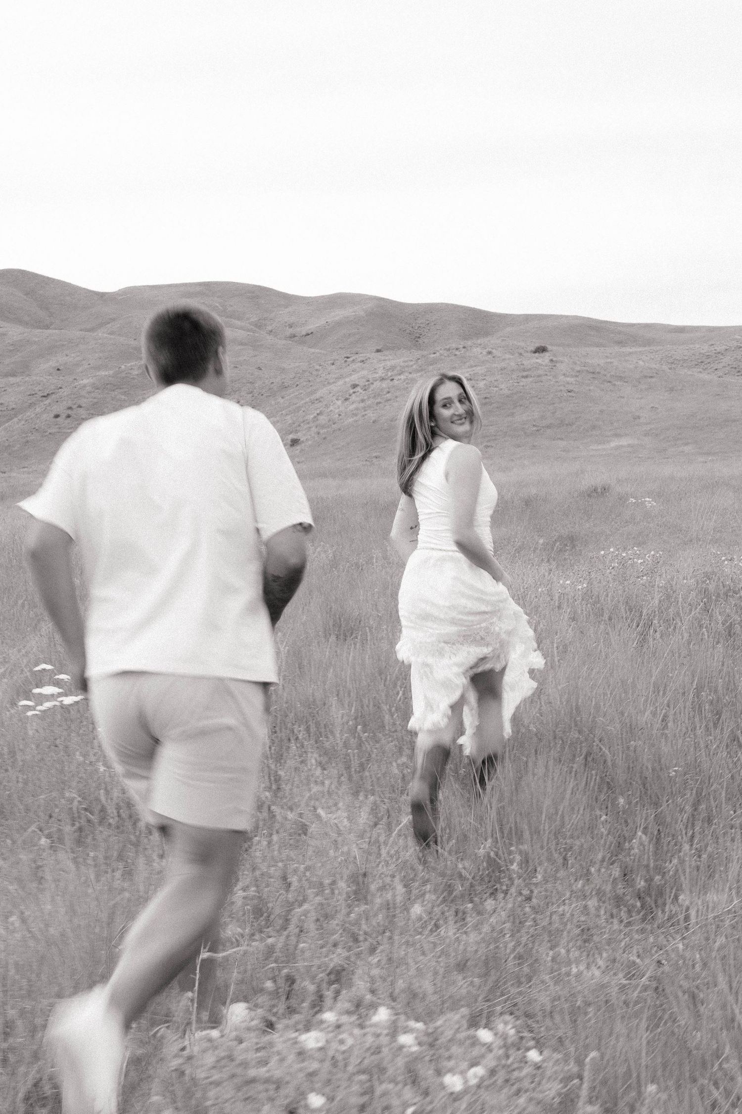 Black and white photo of a couple running through a grassy meadow, woman in white dress smiling back at man.