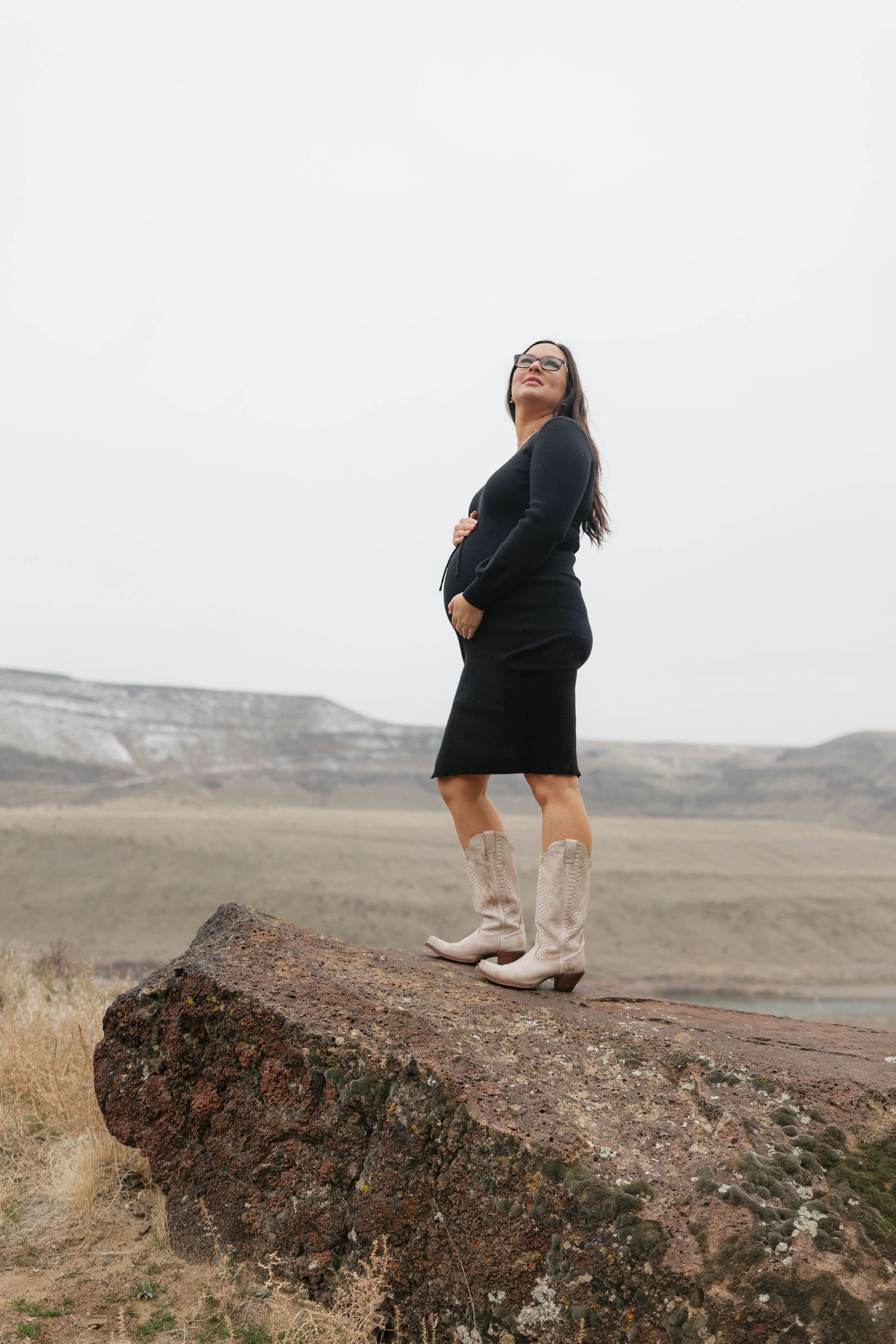 A pregnant woman in a black dress and cowboy boots stands on a rock formation against a cloudy rural landscape.