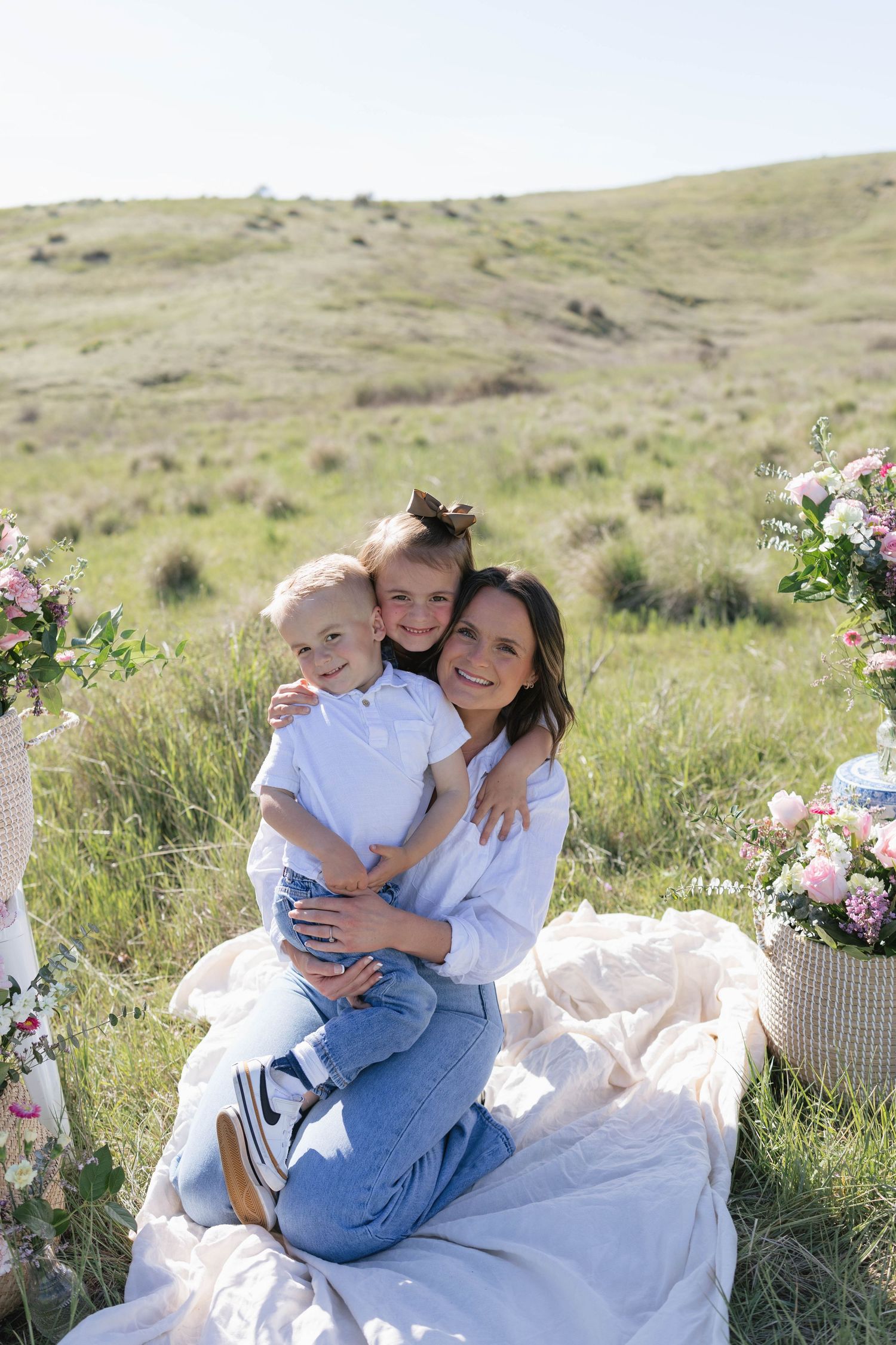 A smiling mother sits on a blanket in a green field, hugging her two young children surrounded by pink floral arrangements.