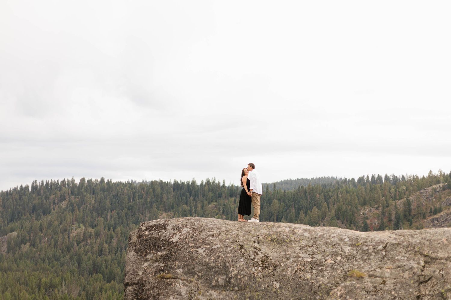 Couple embracing on a rocky cliff overlooking a dense pine forest under an overcast sky.