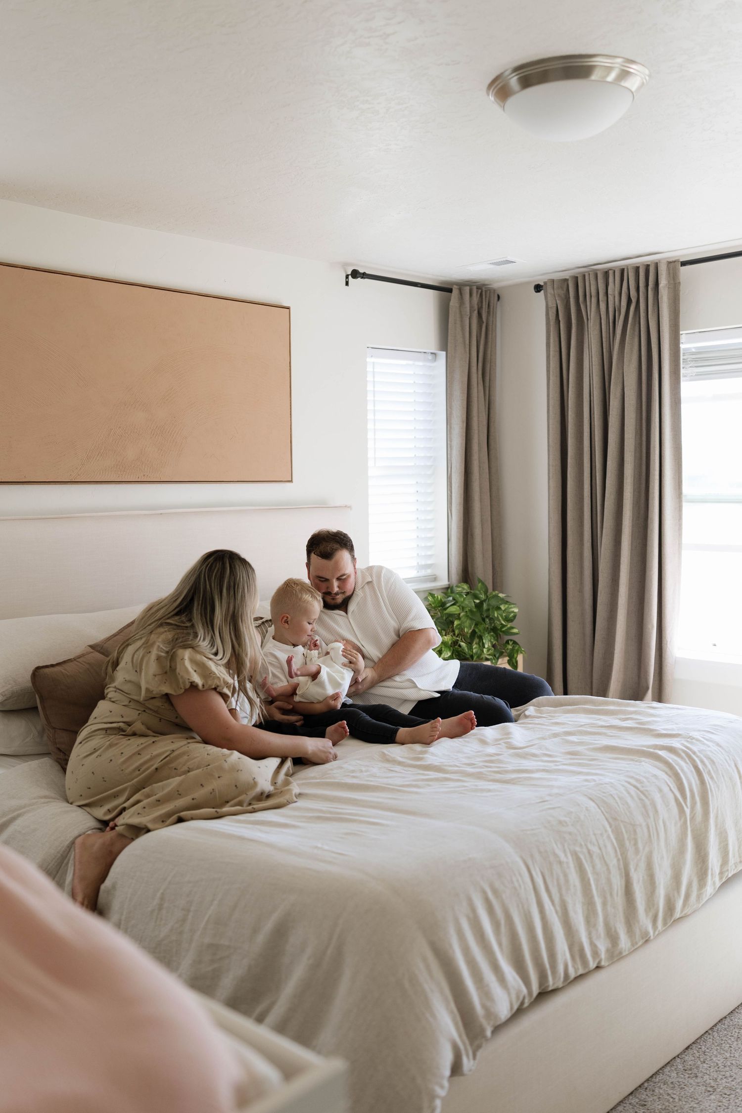 A mother, father, and baby sit together on a neutral linen-covered bed in a bright, cozy bedroom with taupe curtains.