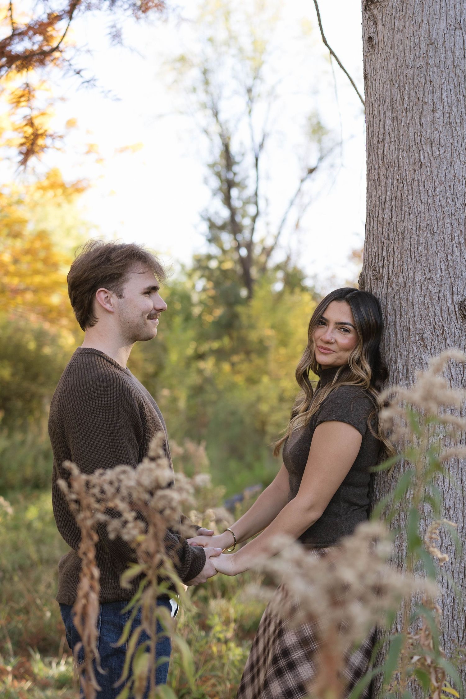 Smiling couple holding hands beside a large tree in an autumn park, surrounded by golden foliage and tall dried wildflowers.