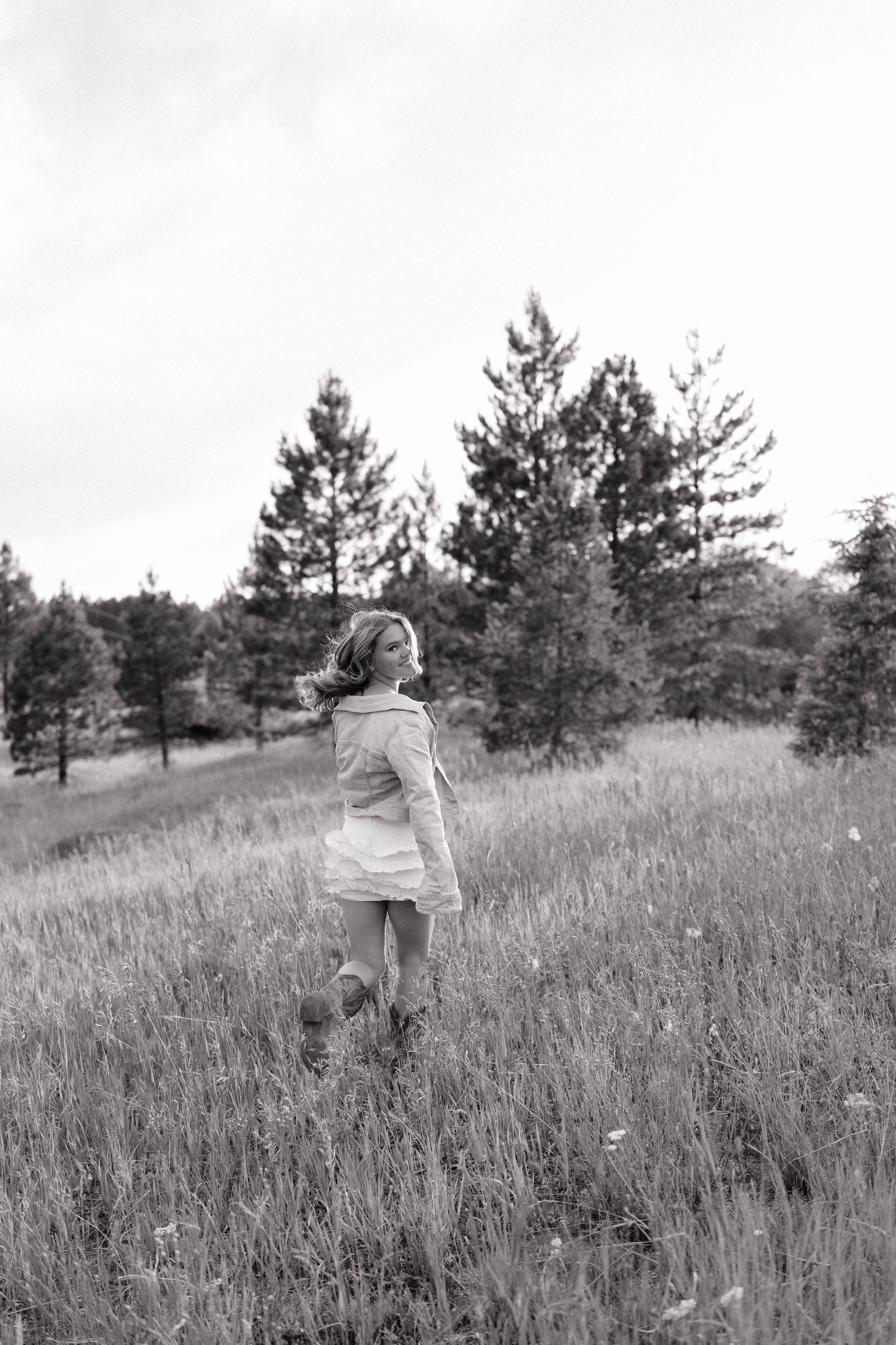 Black and white photo of a young girl walking through a grassy meadow with pine trees in the background.