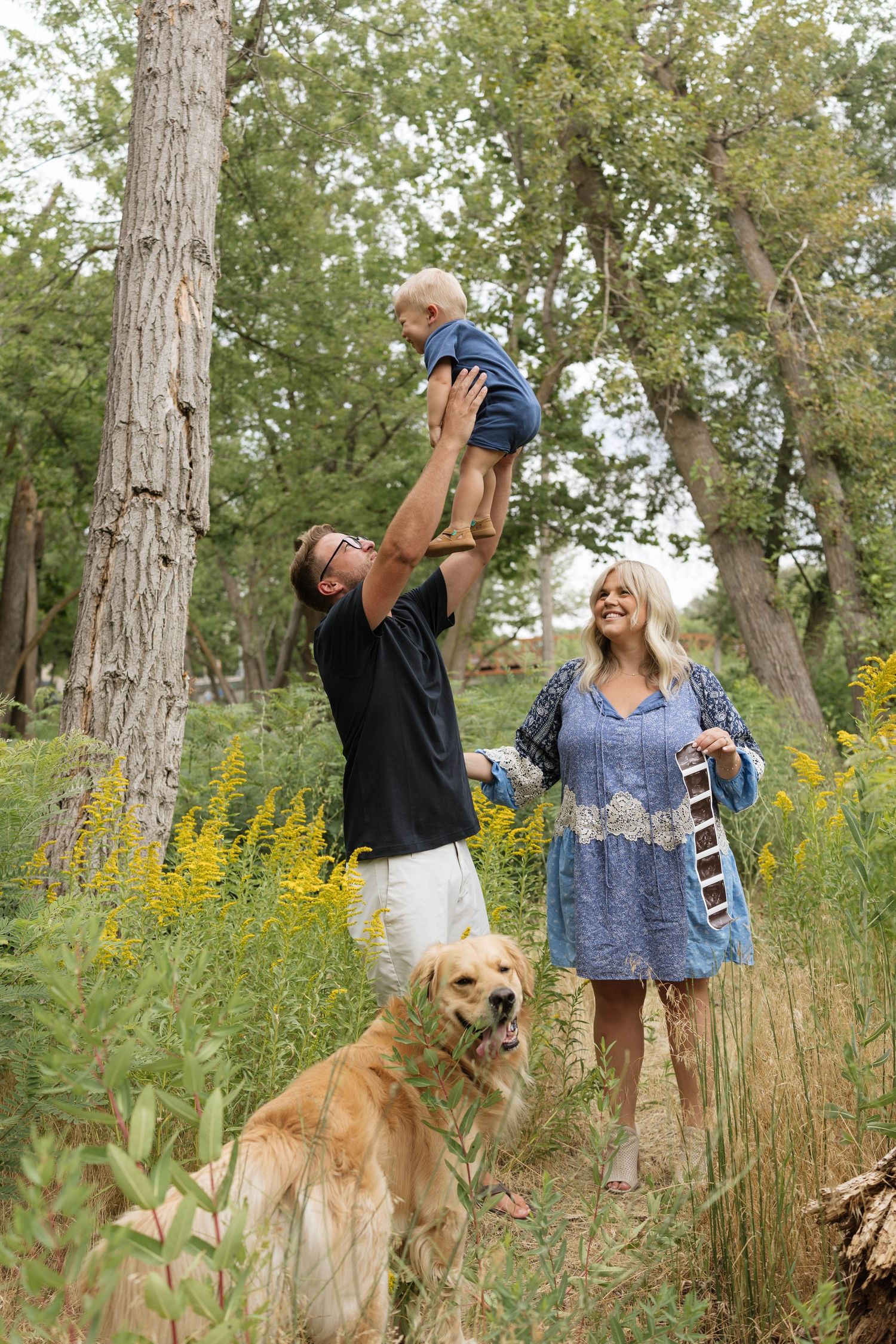 Family with golden retriever in forest, father lifting baby, mother in blue dress smiling outdoors.
