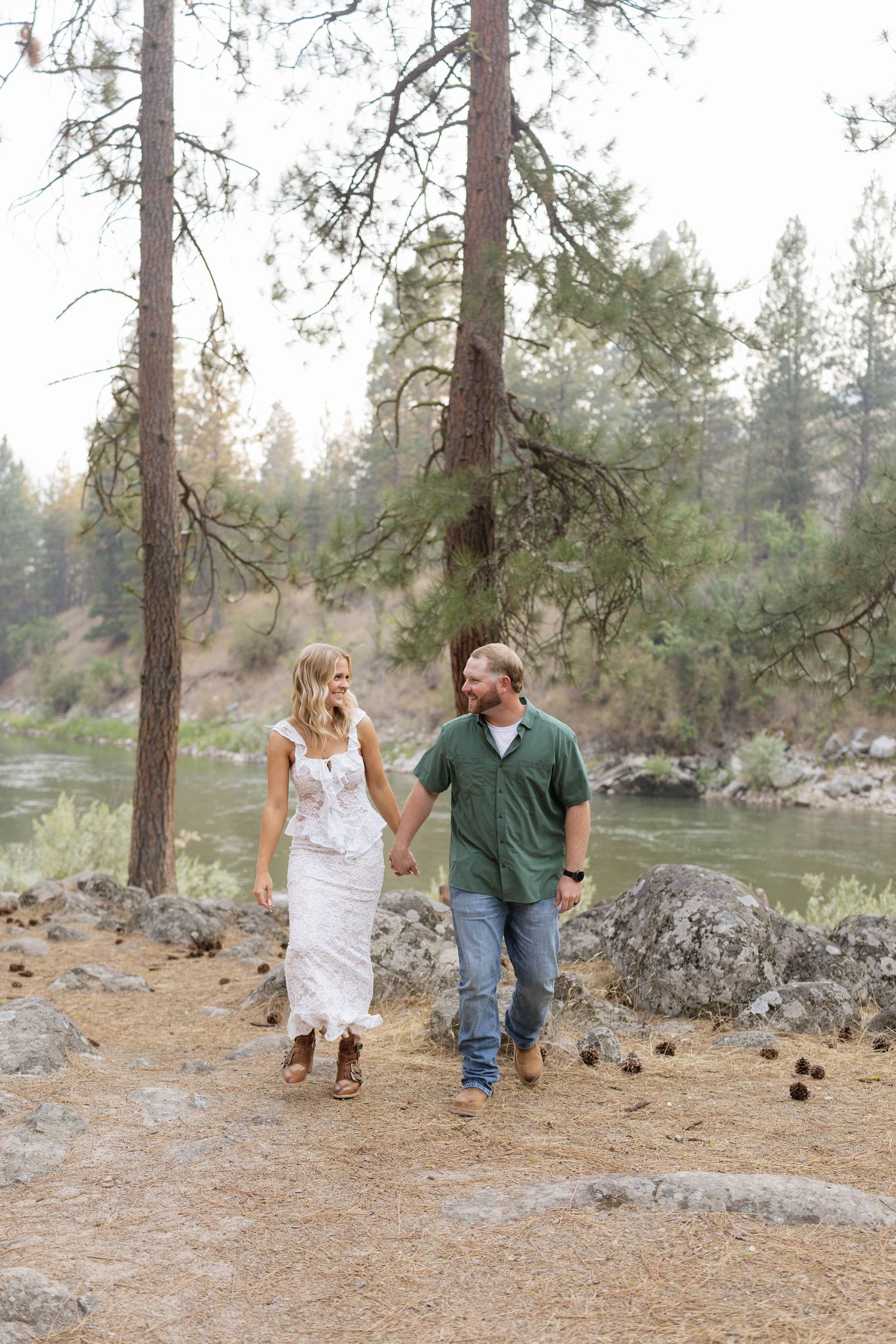 Smiling couple holding hands and walking along a rocky riverbank surrounded by tall pine trees during an outdoor engagement photo session.