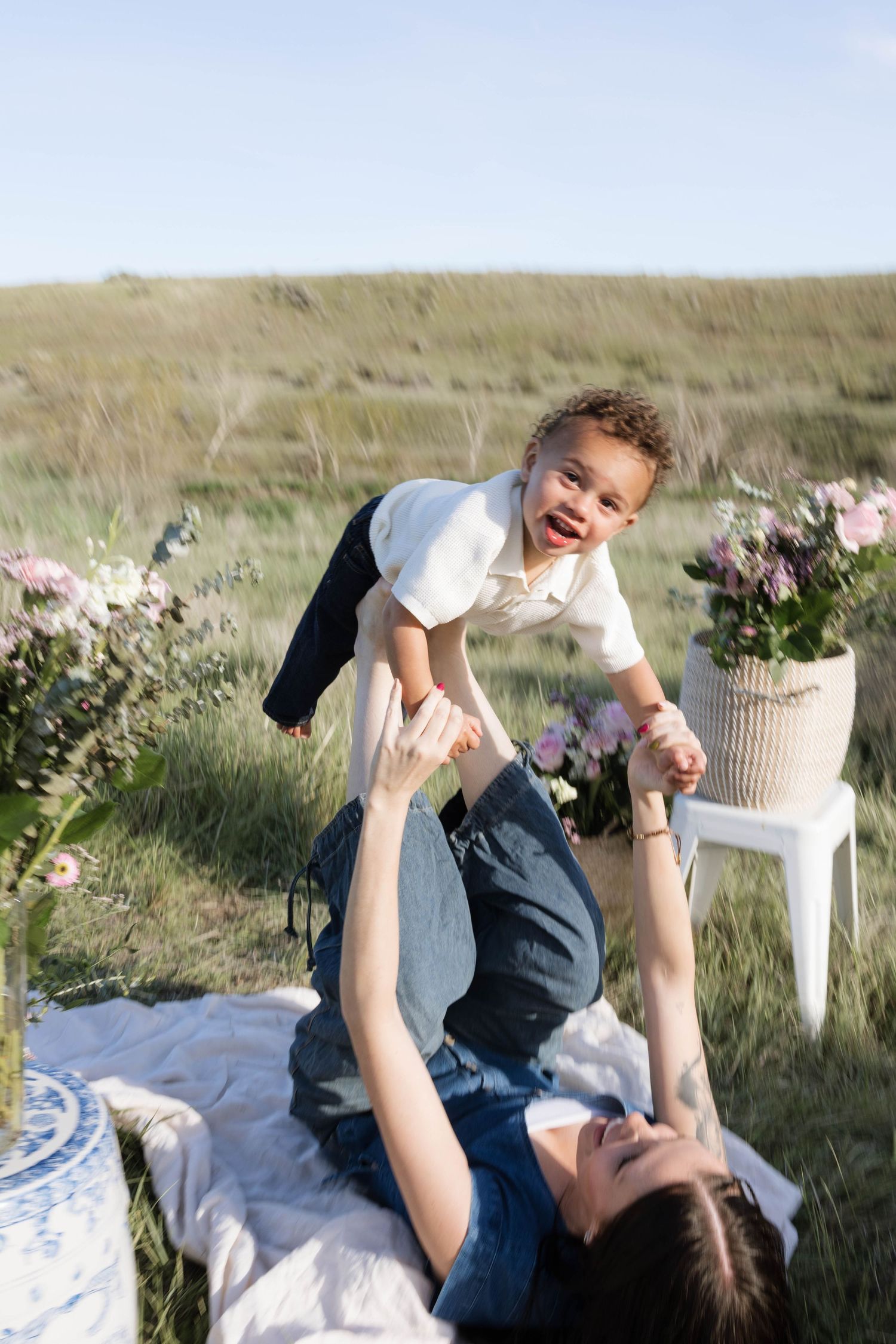A mother lies on a blanket in a field, joyfully lifting her laughing toddler above her during an outdoor picnic.