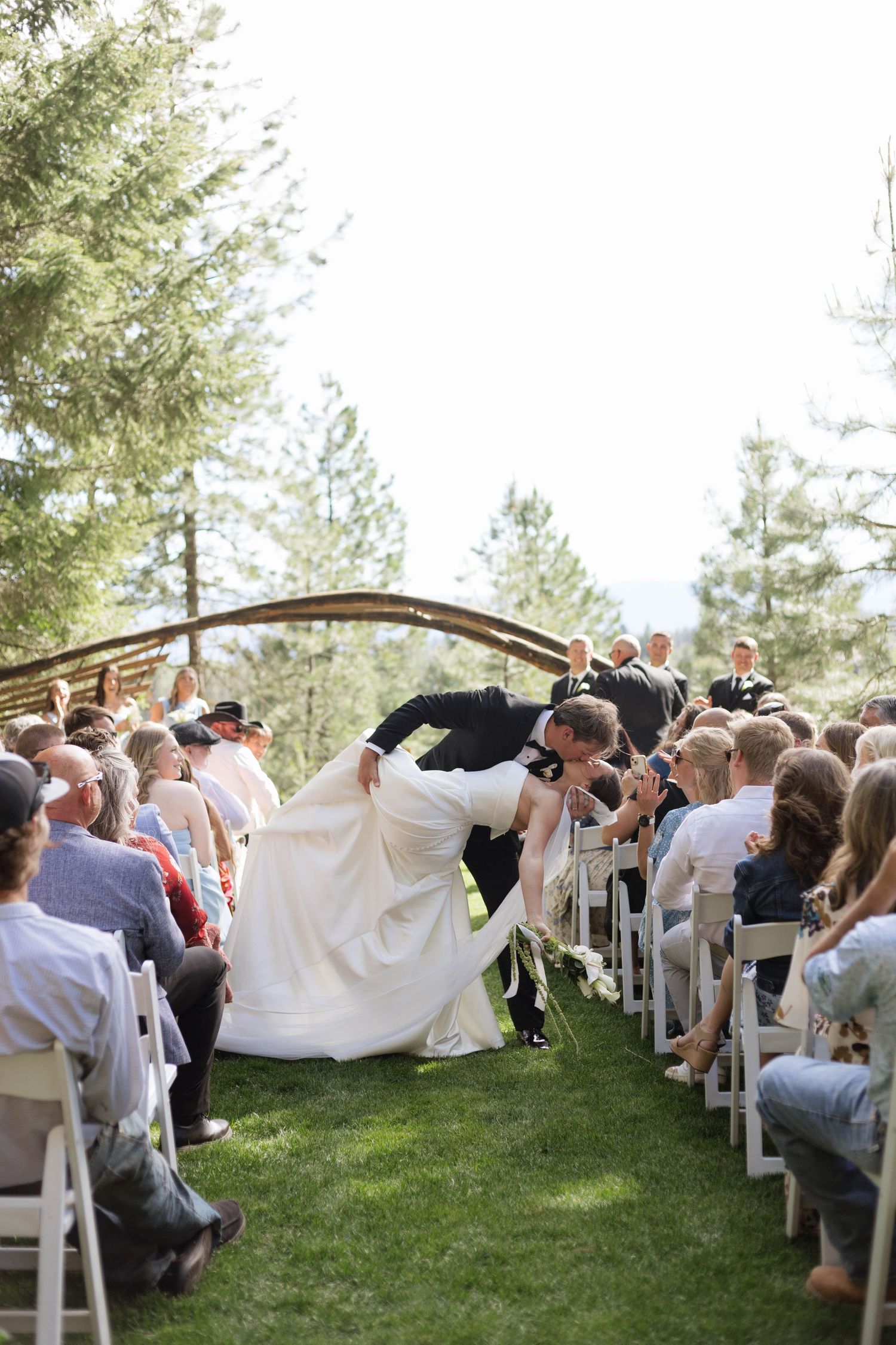 Groom dips bride in wedding aisle during outdoor ceremony with guests seated on white chairs amid pine trees.
