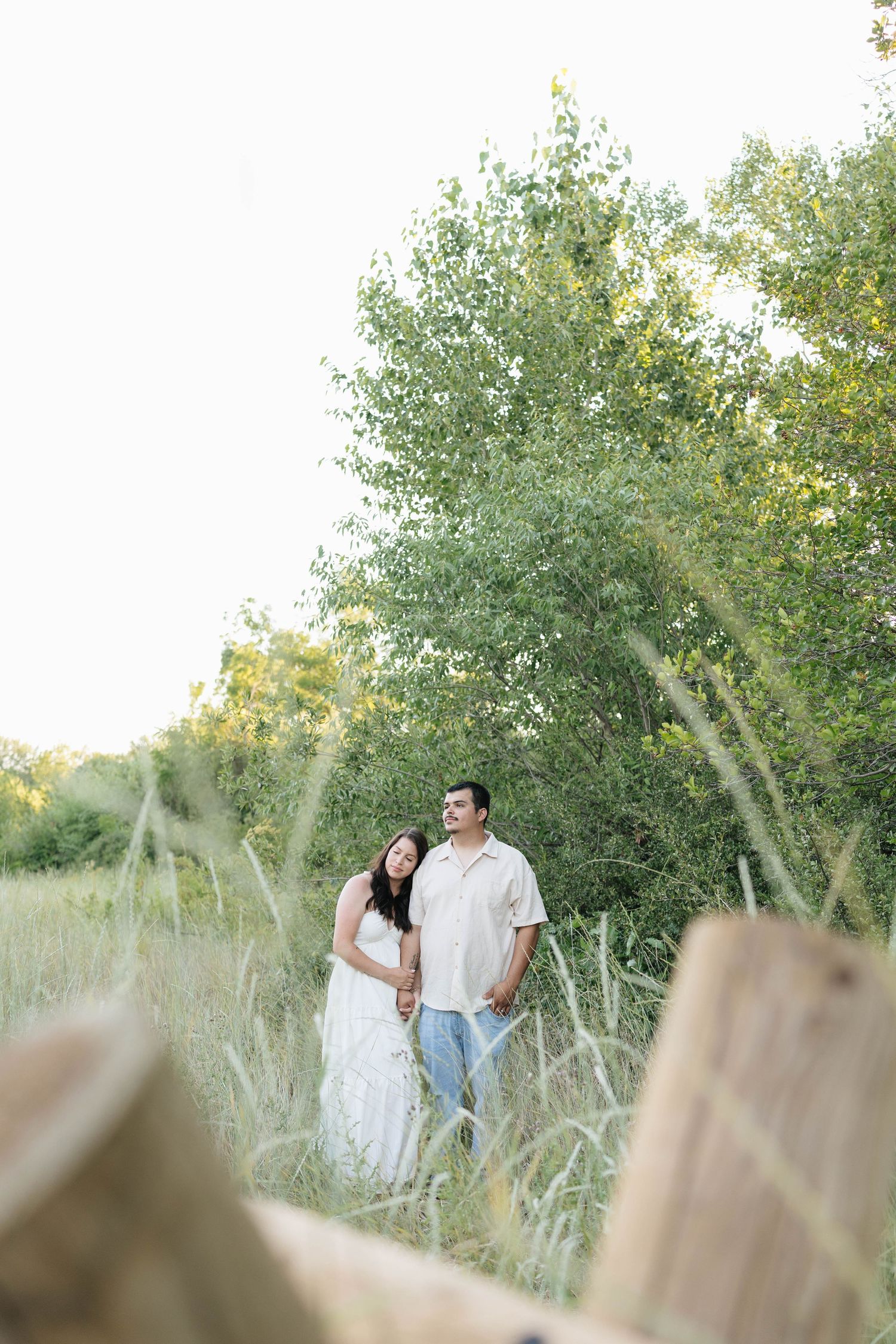 Couple standing together in a lush green meadow near trees, woman in white dress, man in casual attire.